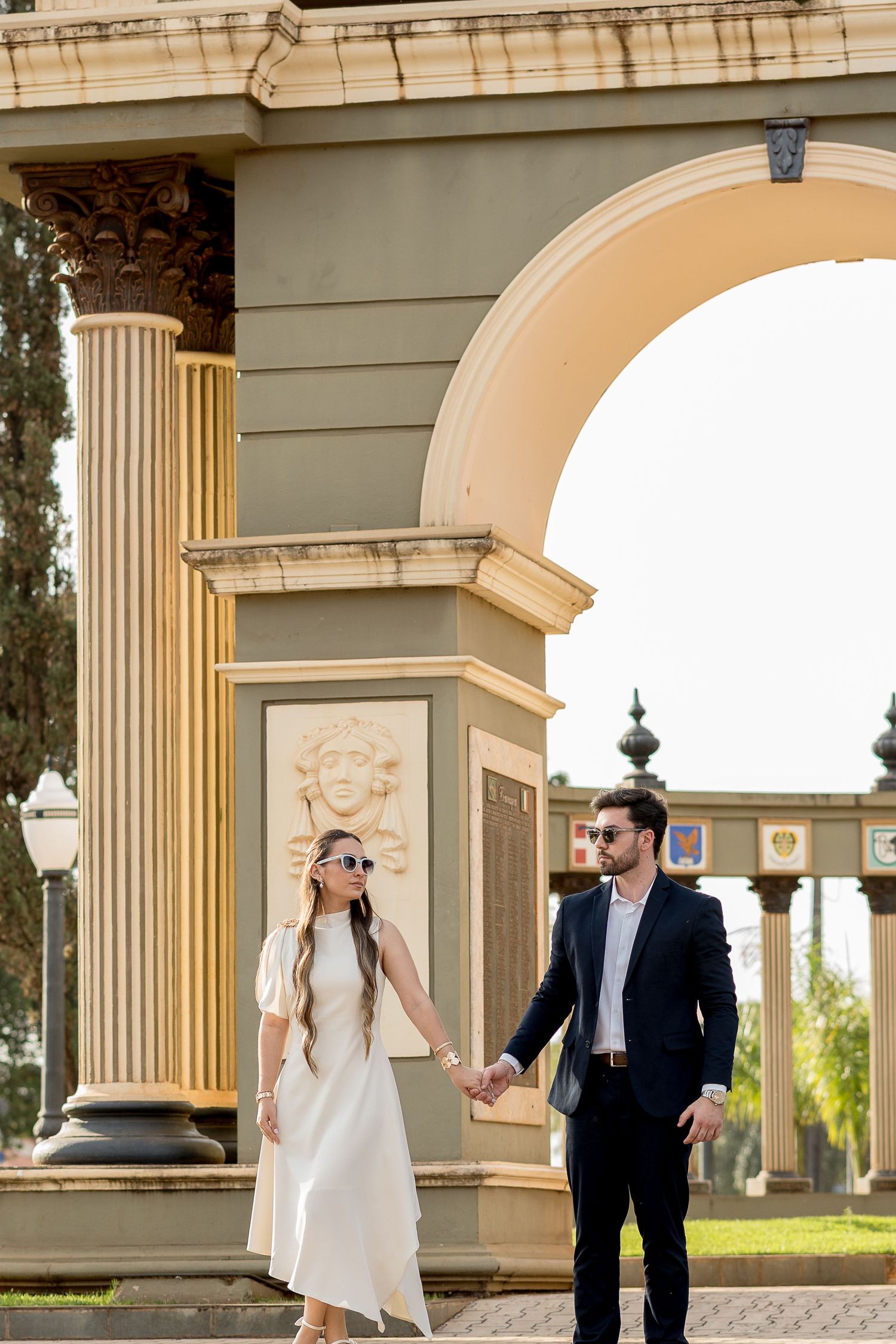 Elegant couple holding hands near ornate arch with columns and relief sculpture in a sunny outdoor setting.