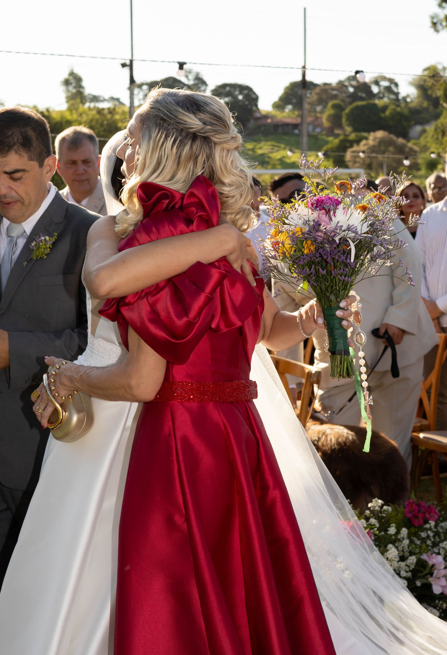 Bride in white gown hugging guest in red satin dress while holding colorful bouquet at outdoor wedding ceremony.