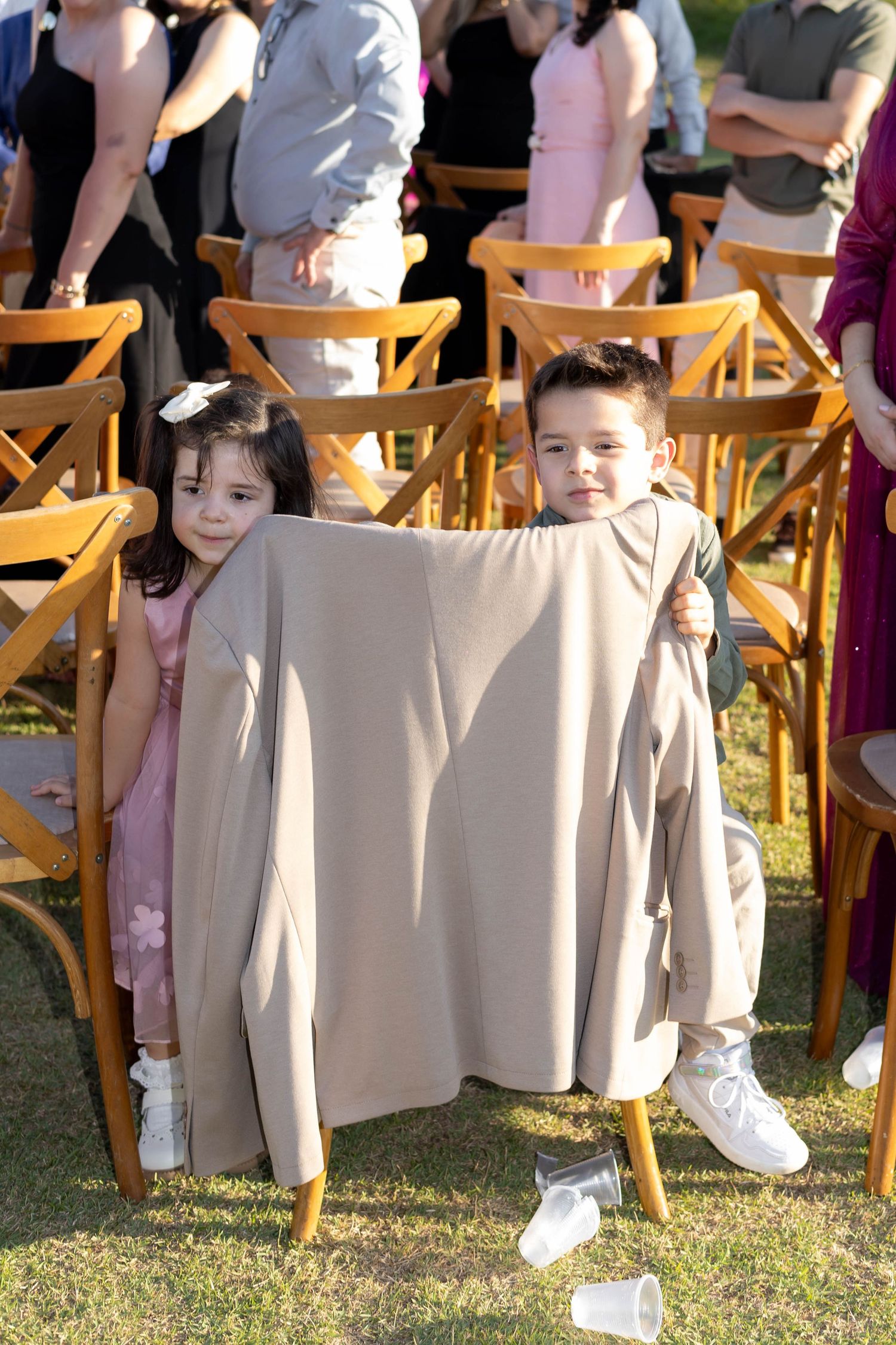 Two children playing behind a beige jacket between wooden chairs at an outdoor wedding ceremony.