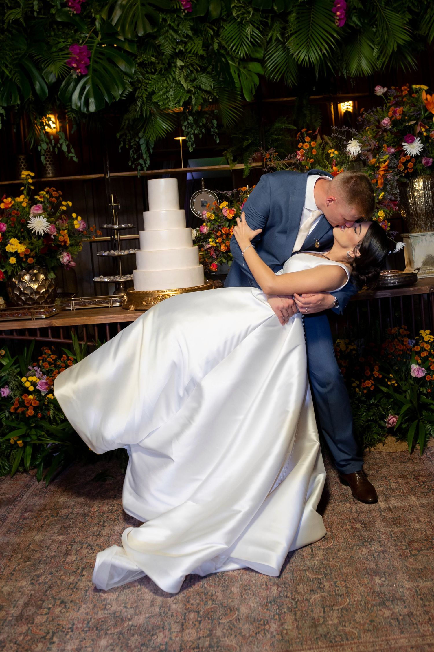 Bride and groom share a romantic dip kiss on the dance floor with a white wedding cake and floral backdrop.