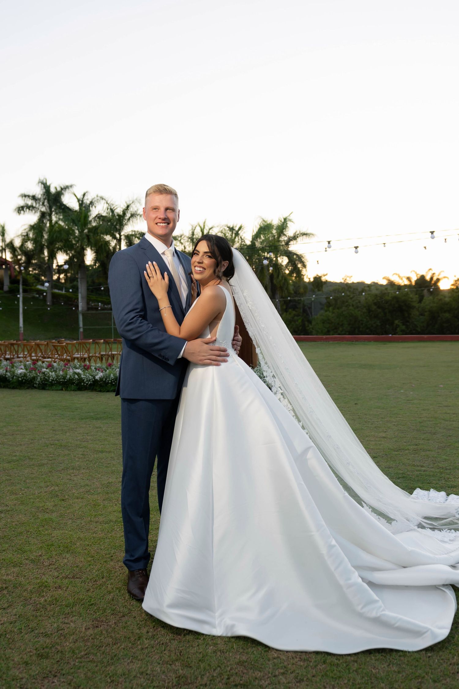 Bride in white ball gown with long veil embraces tall groom in navy suit on green lawn with palm trees.