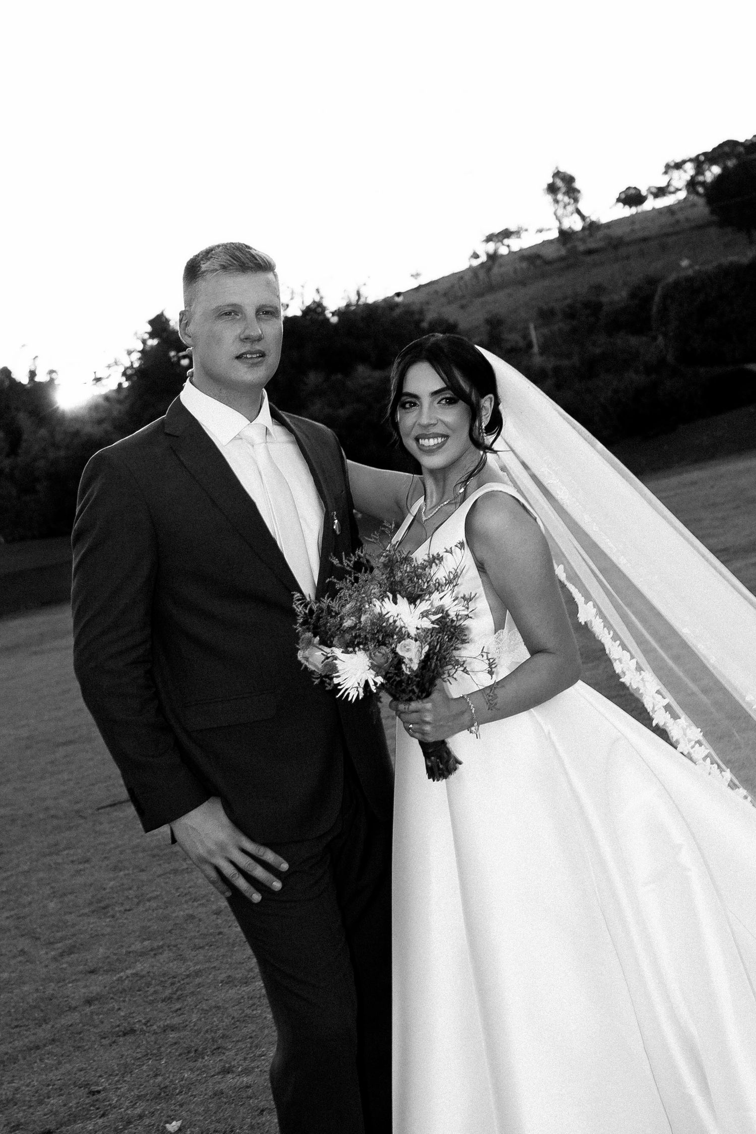 Black and white wedding photo of bride in gown with veil holding bouquet beside groom in suit outdoors.