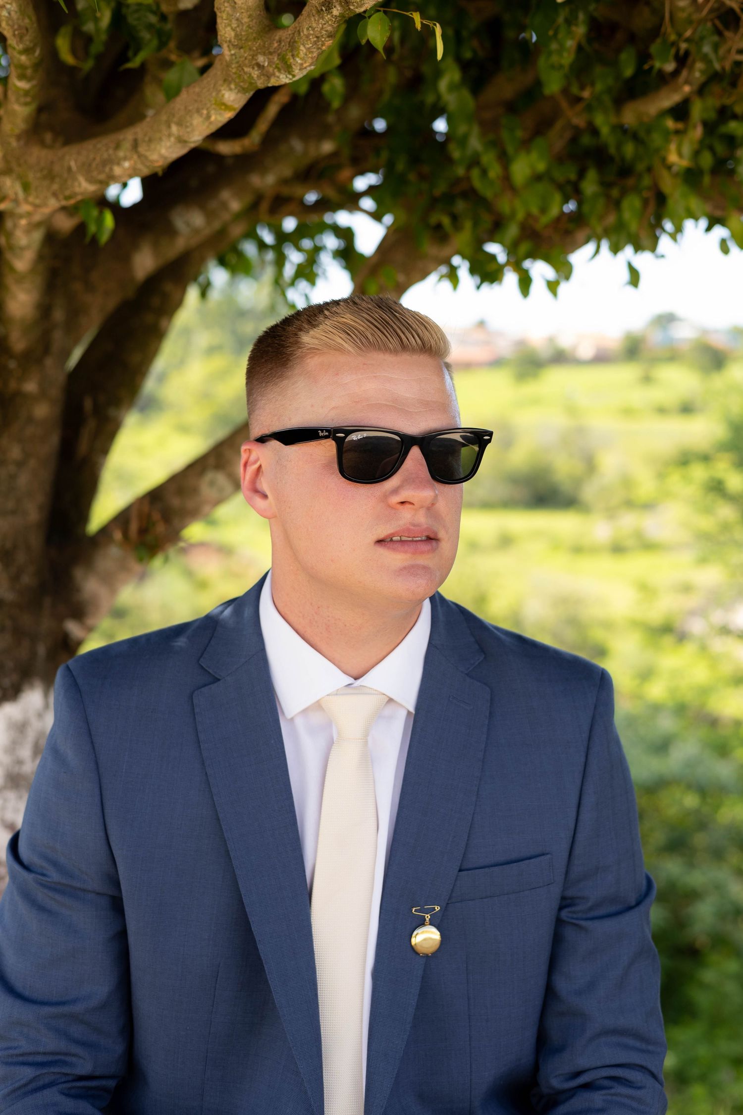 Young man in blue suit, white tie, and black Ray-Ban sunglasses standing under a tree outdoors.