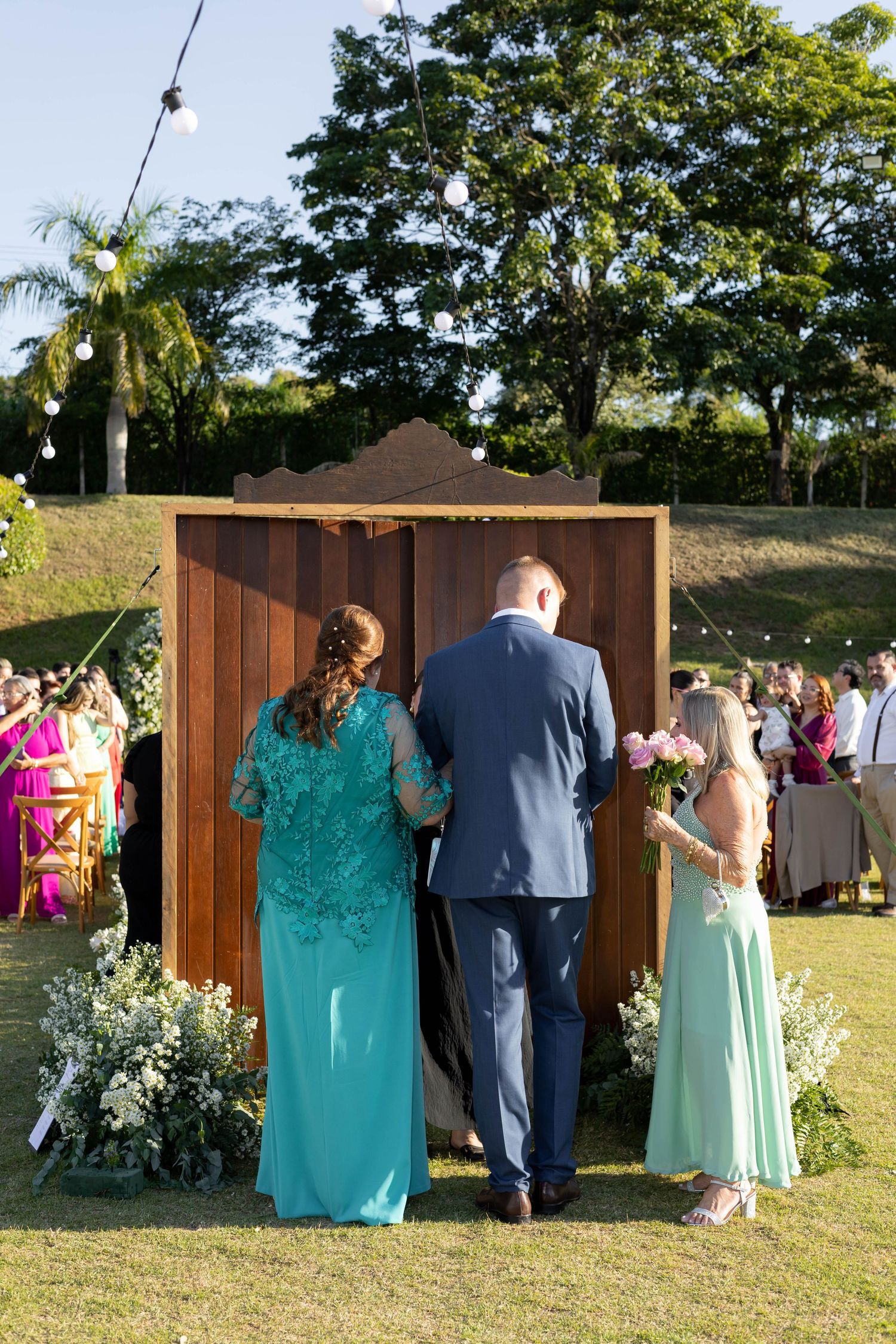 Outdoor wedding ceremony with guests in teal dresses and blue suit standing before a wooden altar surrounded by floral arrangements.