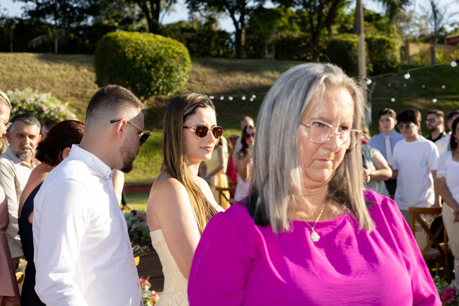 Outdoor wedding gathering with guests in formal attire, woman in pink top in foreground, lush green garden background.