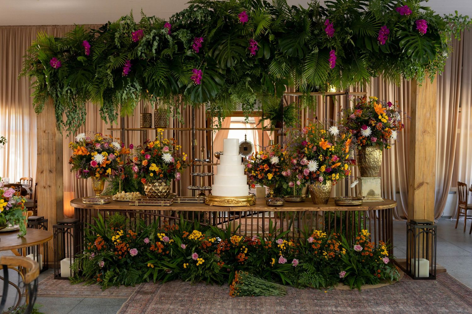 Lush tropical wedding cake table adorned with colorful florals, greenery canopy, and golden lanterns in an elegant venue.