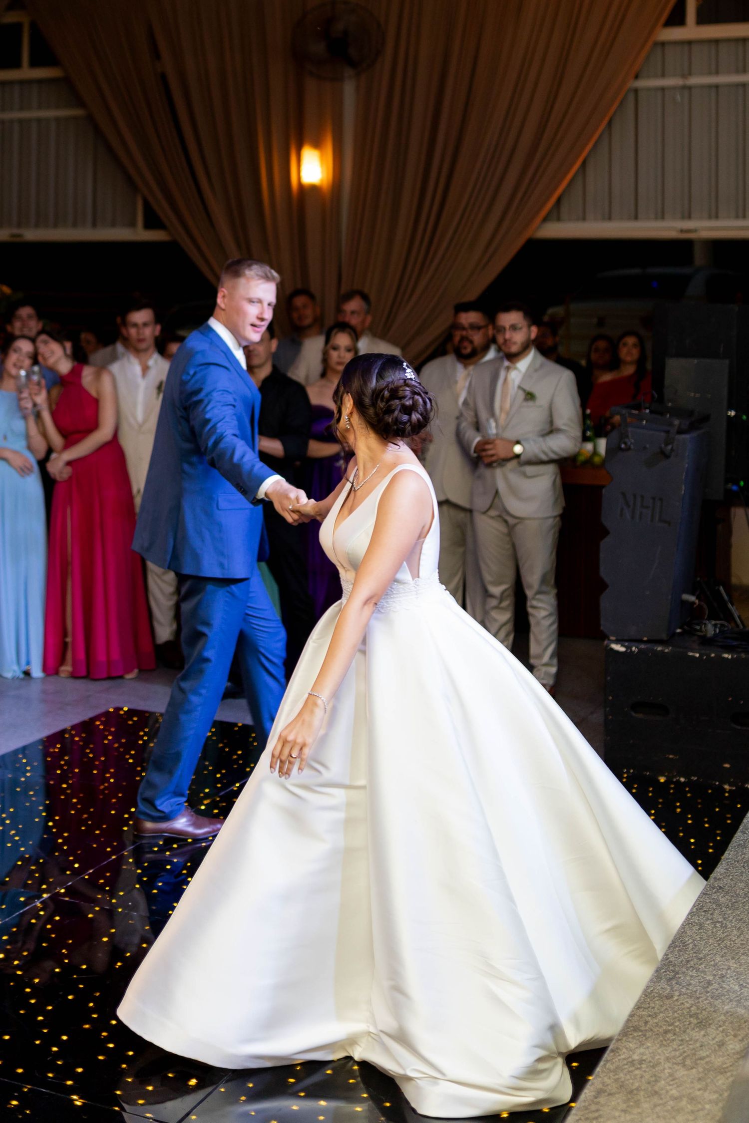 Bride in white ball gown and groom in blue suit share their first dance on a starlit floor at wedding reception.