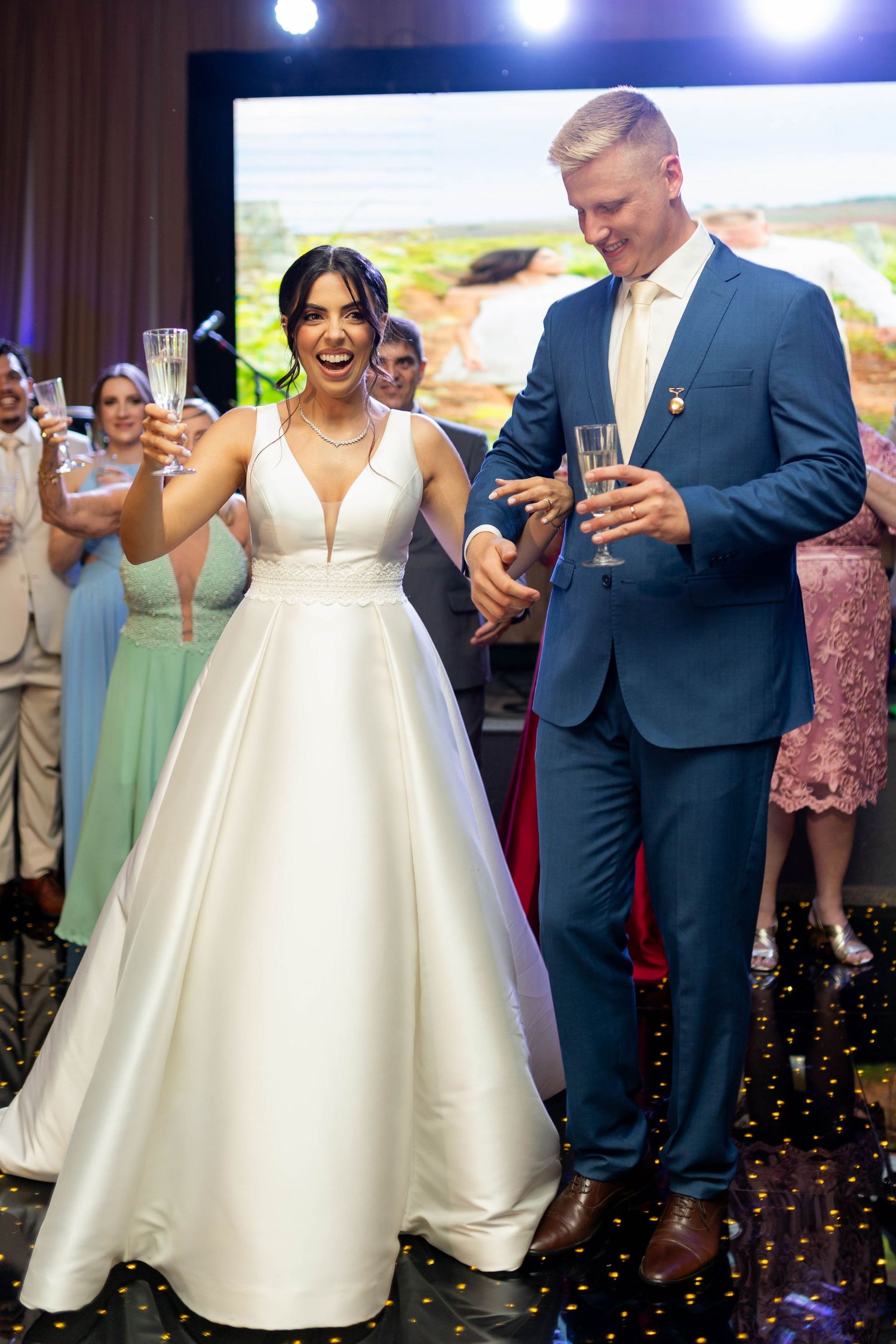 Joyful bride in white gown and groom in blue suit toasting champagne at their wedding reception.