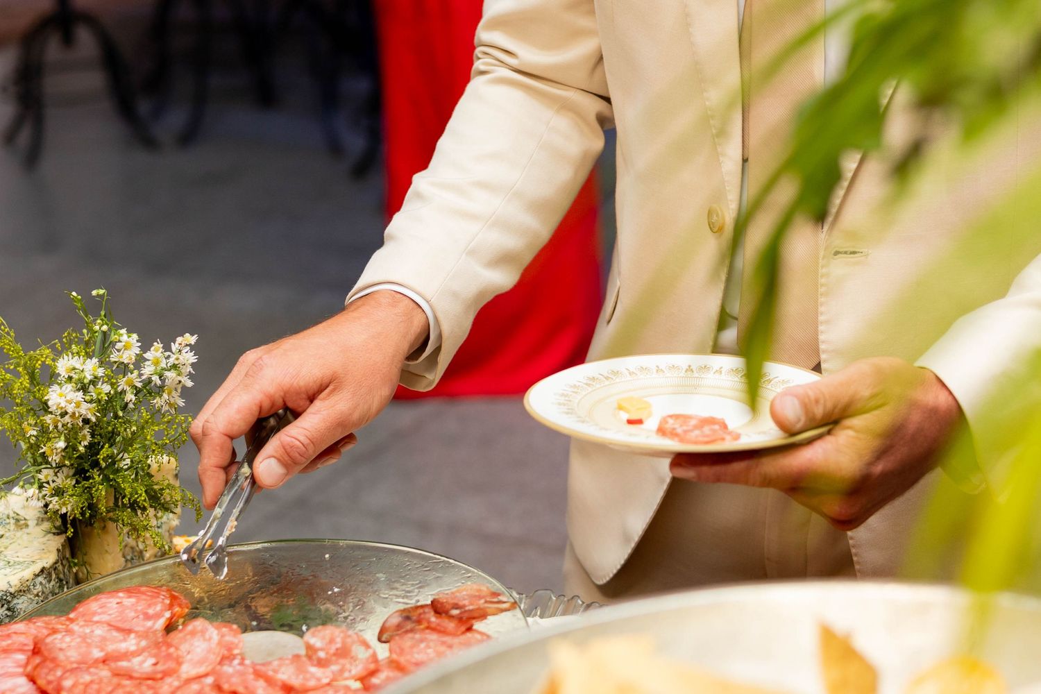 Person serving charcuterie at a buffet table, holding a white plate while using tongs to select cured meats.
