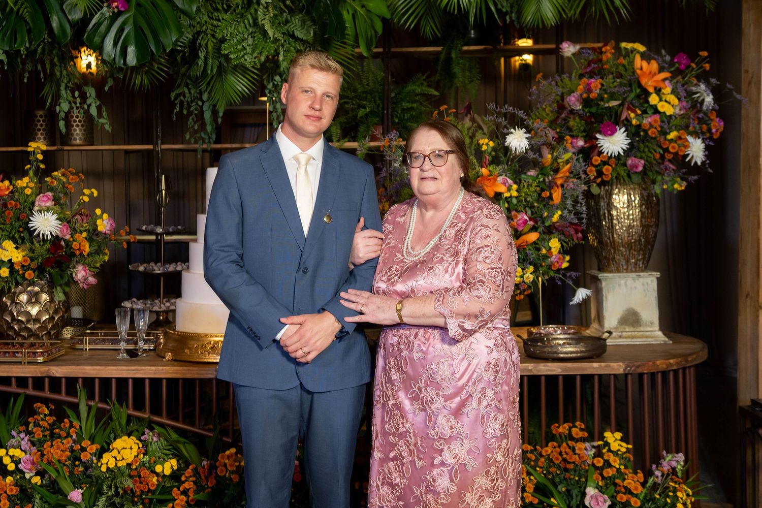 A man in a blue suit and woman in pink lace dress pose together among lush floral arrangements at an elegant event.