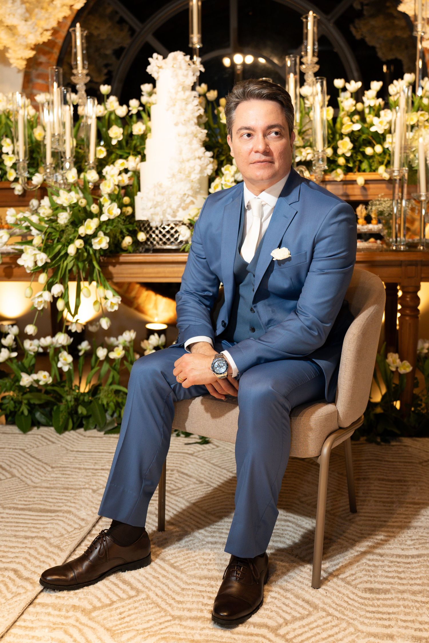 Man in blue three-piece suit sitting elegantly at a floral wedding reception with white flowers and cake backdrop.