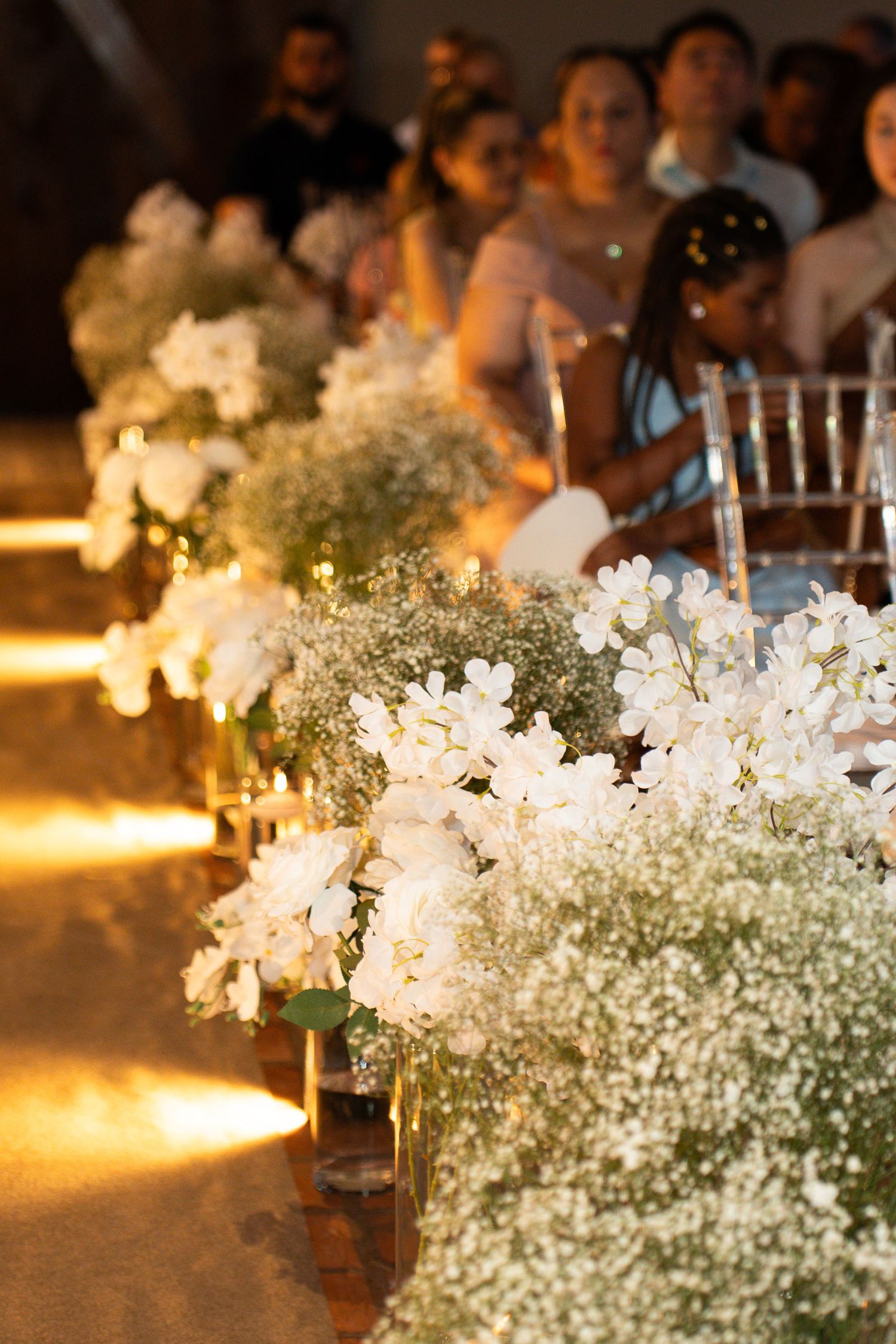 White orchids and baby's breath floral arrangements line a wedding aisle with warm golden lighting and seated guests.
