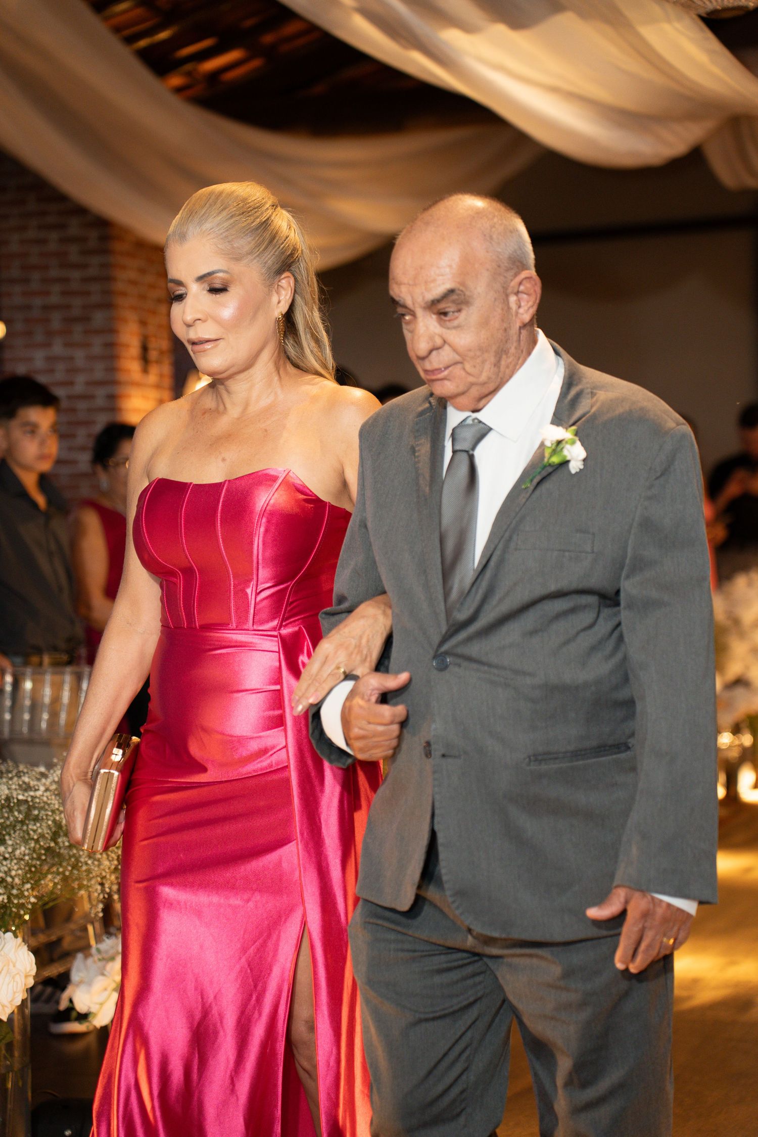 Elegant couple at a wedding, woman in red satin gown and man in gray suit with boutonniere, walking arm in arm.