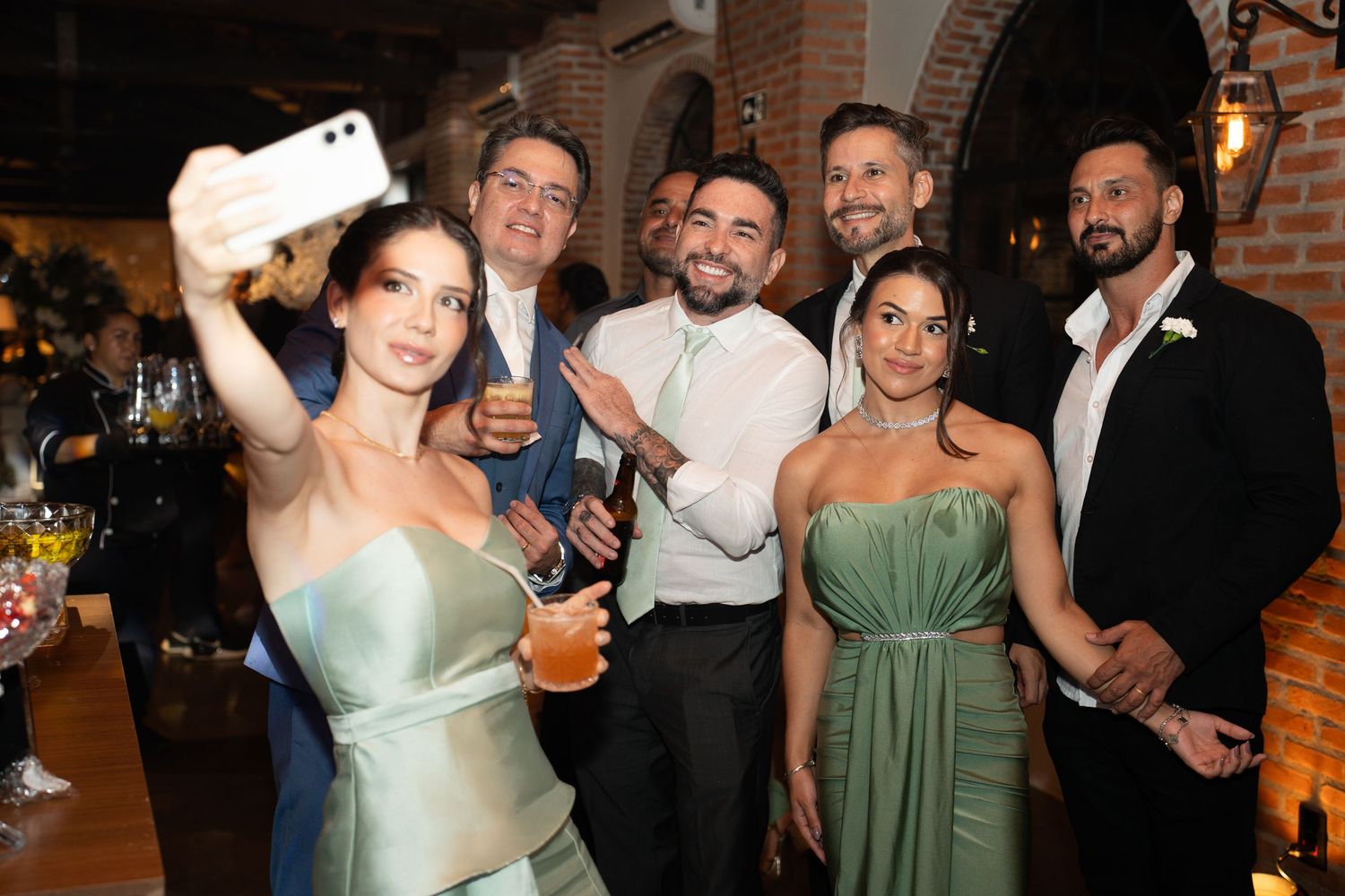Group of elegantly dressed guests taking a selfie at a wedding reception in a warmly lit venue.