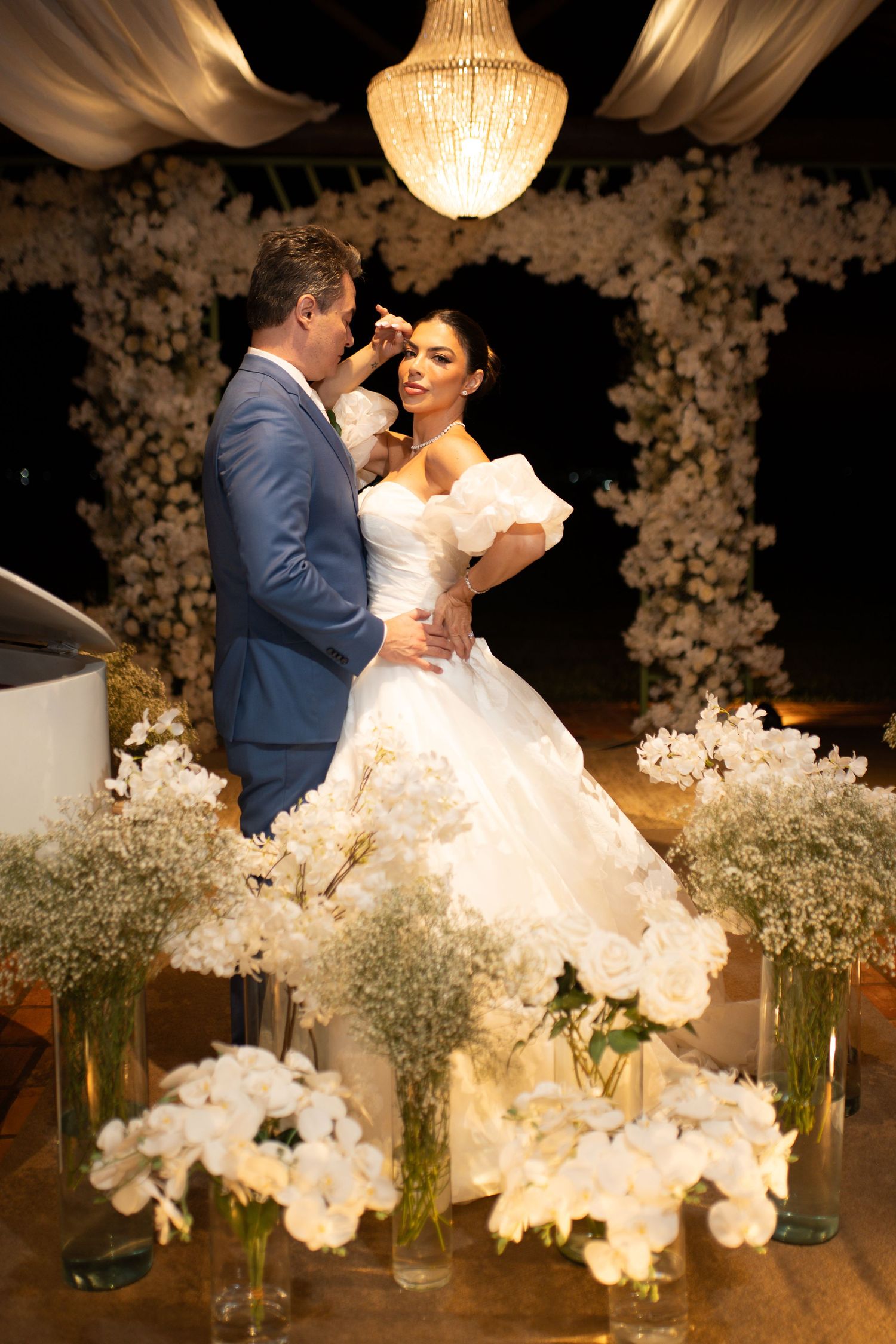 Bride and groom share a romantic moment at elegant night wedding with white floral arrangements and chandelier backdrop.
