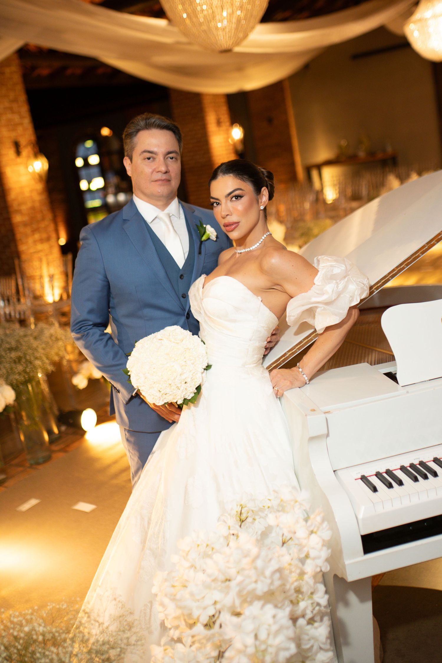 Elegant bride in white gown with bouquet and groom in blue suit pose by a white grand piano at wedding reception.