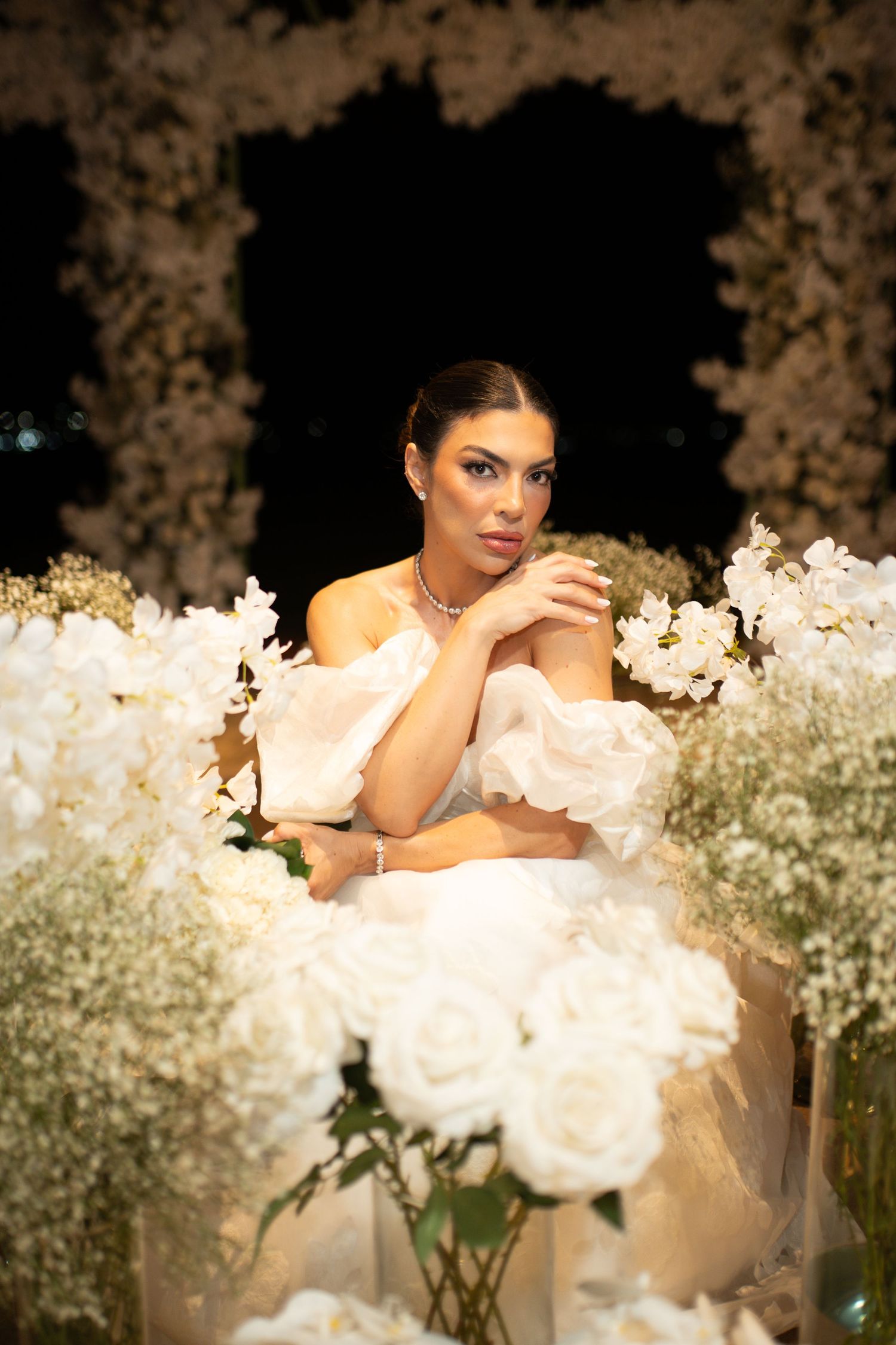 Elegant bride in white off-shoulder gown surrounded by lush white roses and floral arrangements in a romantic night setting.