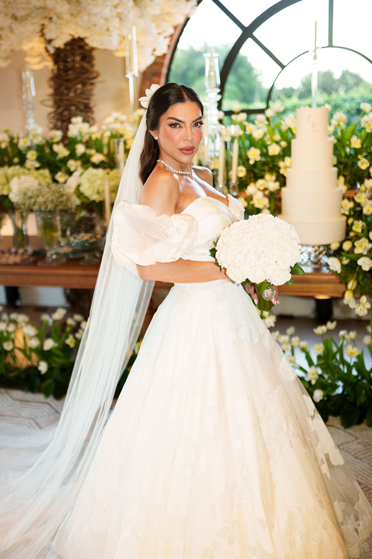 Bride in white off-shoulder ball gown with veil holds hydrangea bouquet amid floral wedding decor.