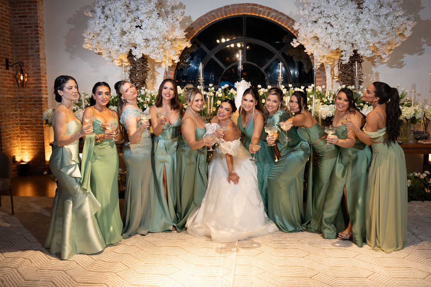 Bride in white gown poses with bridesmaids in sage green dresses at elegant floral wedding reception.