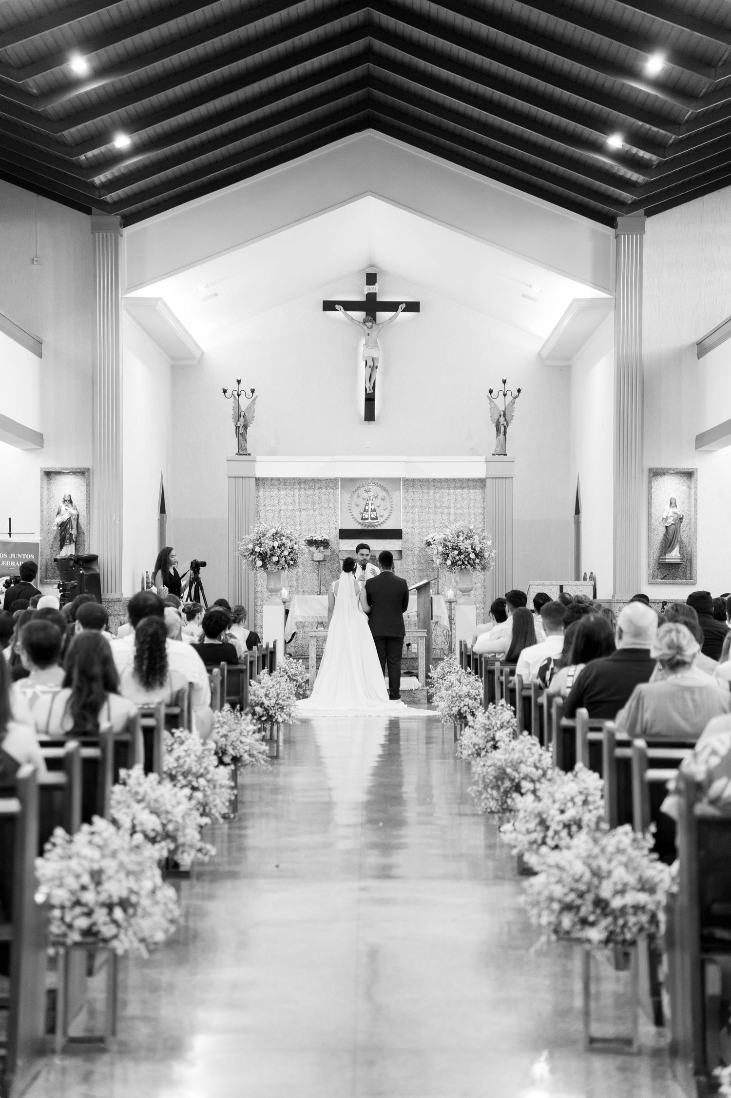 Black and white photo of a couple exchanging vows at a Catholic church wedding ceremony with floral aisle decor.