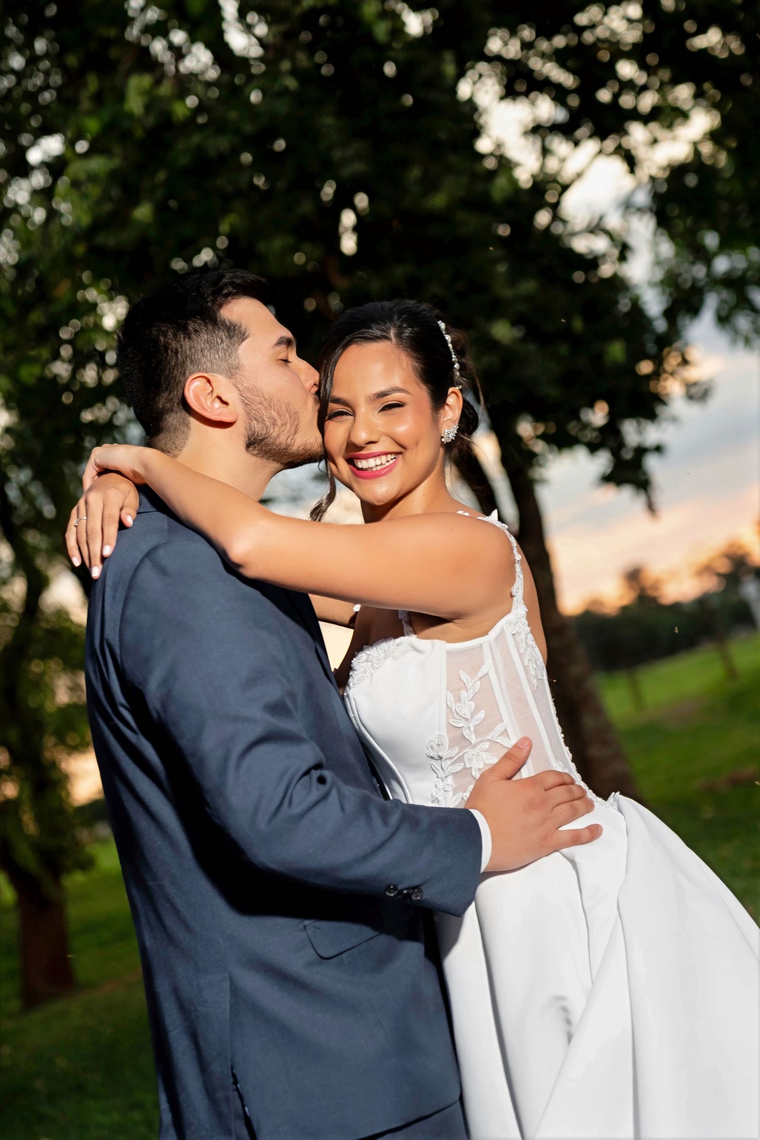 Bride in white lace gown smiles as groom in navy suit kisses her cheek outdoors at sunset.