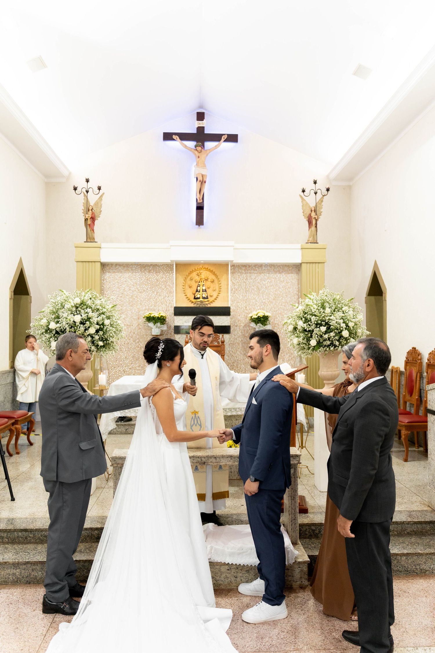 Bride and groom exchange vows at a Catholic church altar, flanked by family members, with a crucifix above.