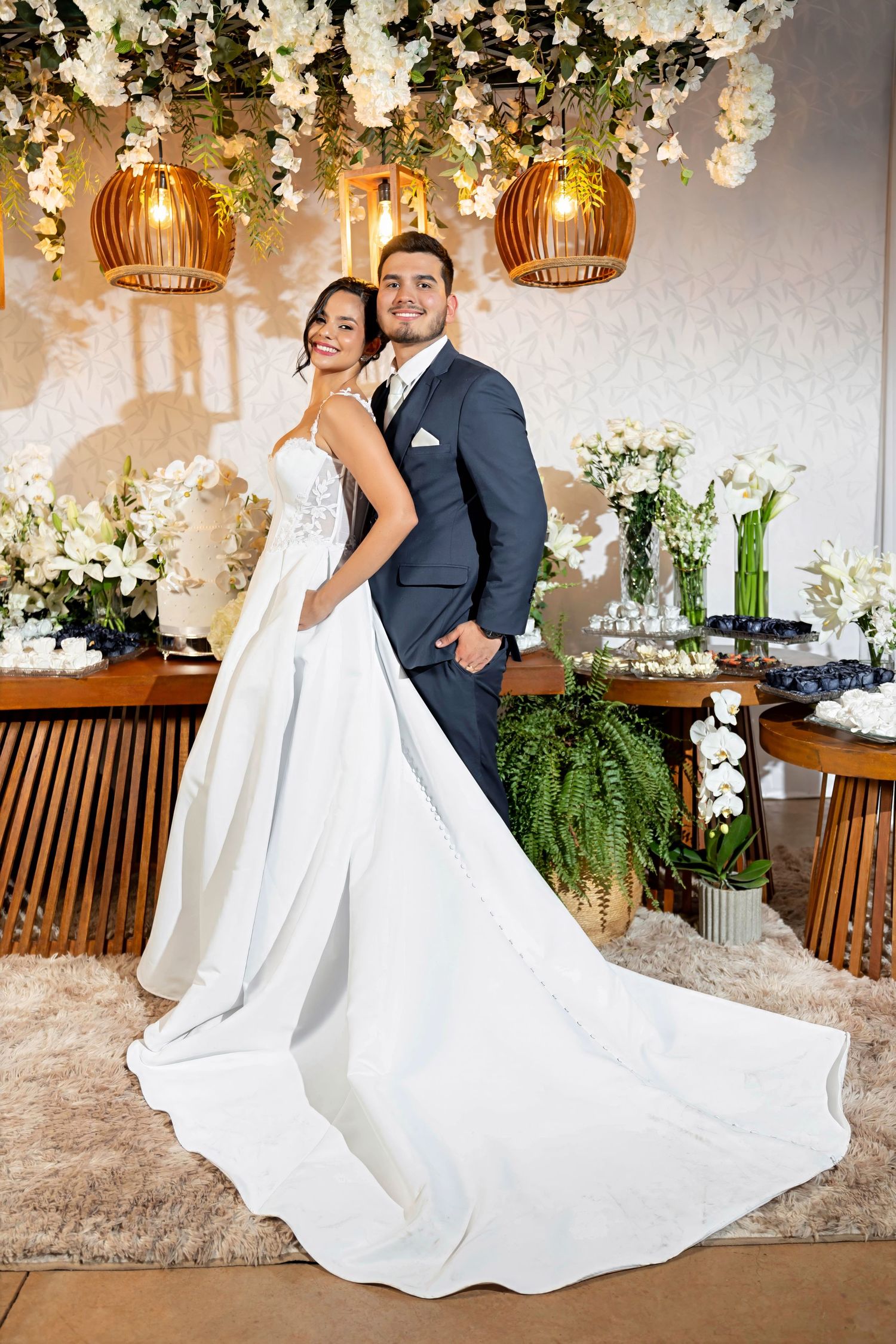 Bride in white gown and groom in navy suit pose elegantly at floral wedding reception decor.