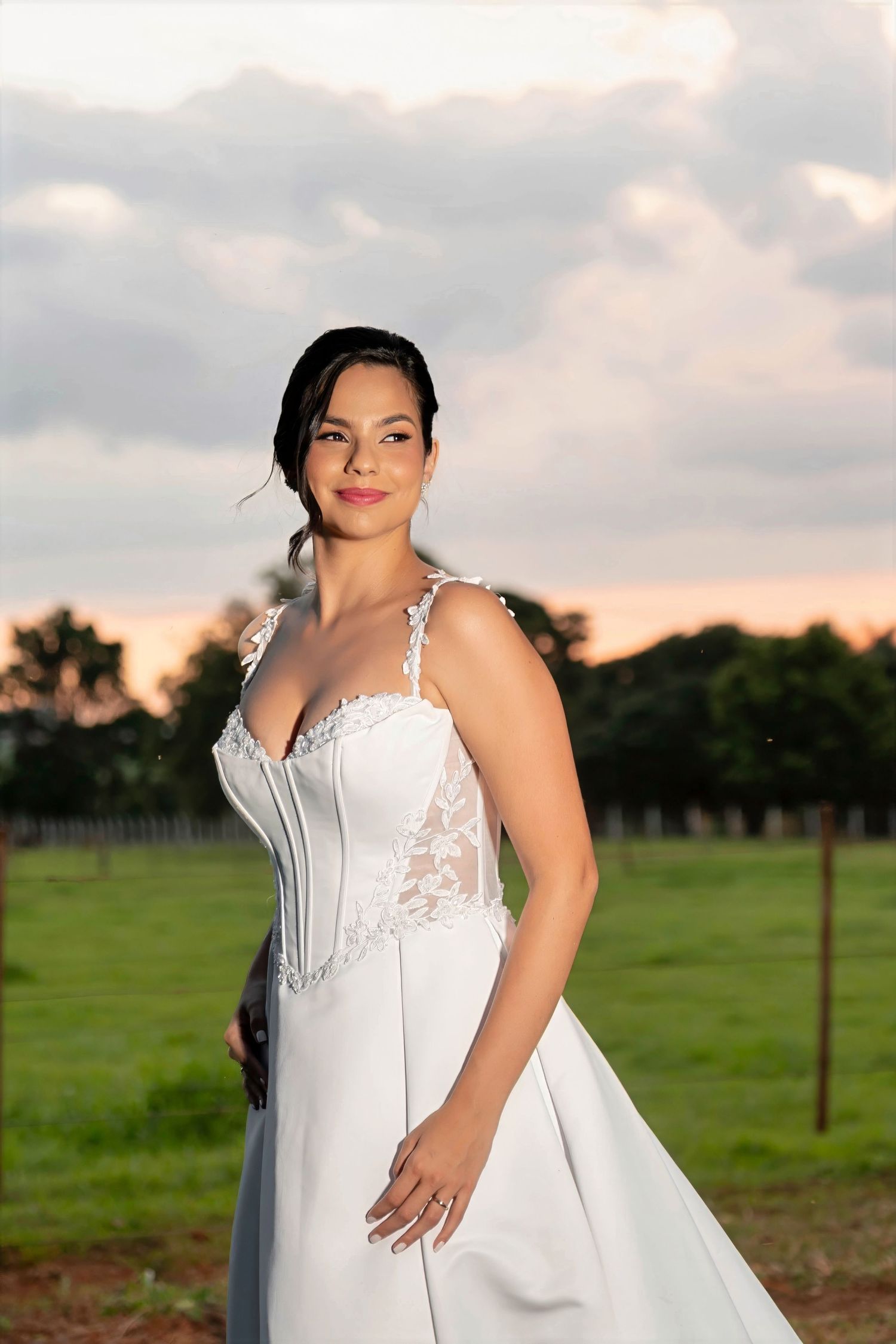 Bride in white lace corset wedding gown poses outdoors at sunset with green fields and cloudy sky.