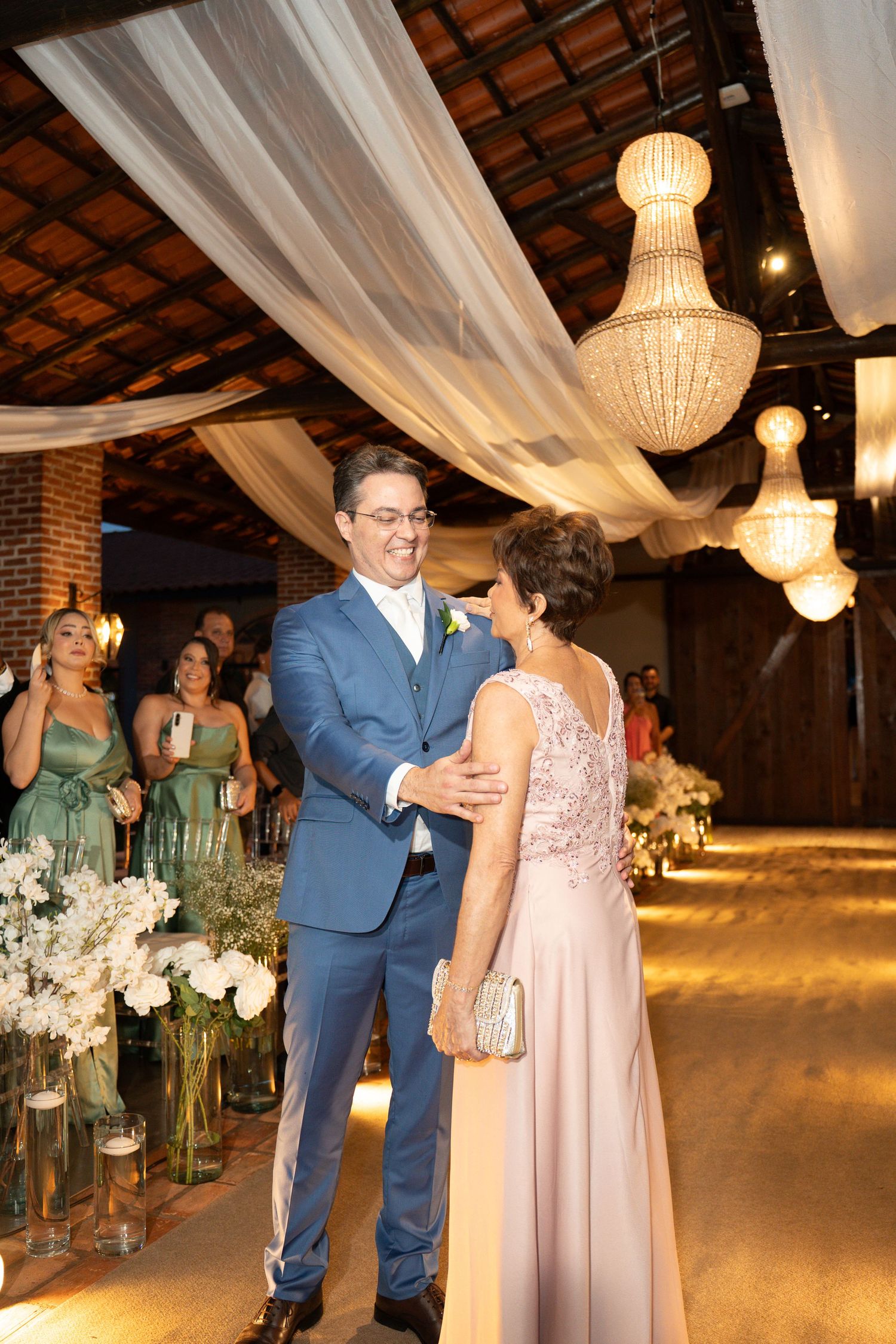 Groom in blue suit dancing with woman in pink gown at elegant wedding reception with chandeliers.