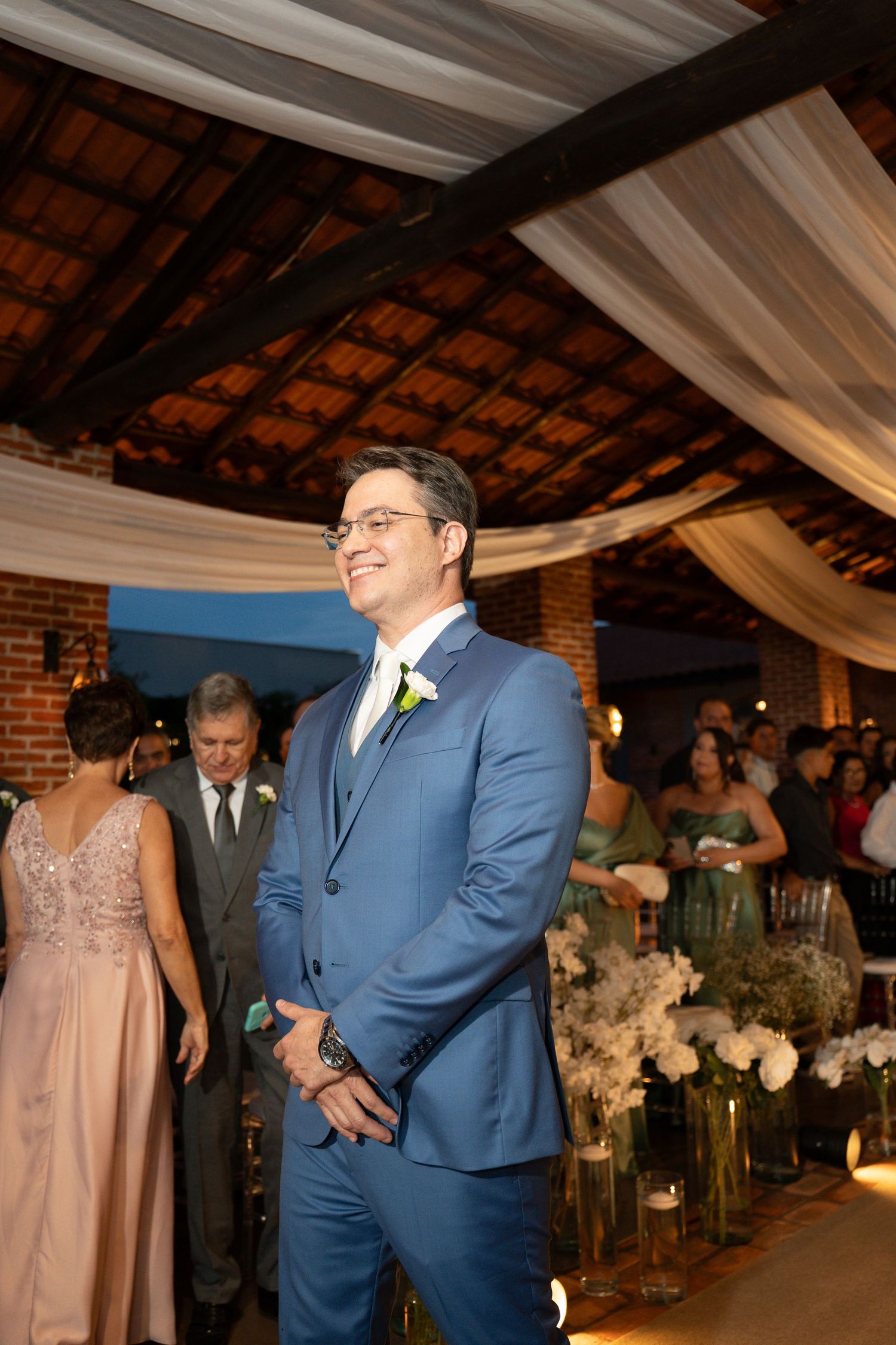 Groom in blue suit smiling at wedding ceremony aisle with floral arrangements and guests in background.