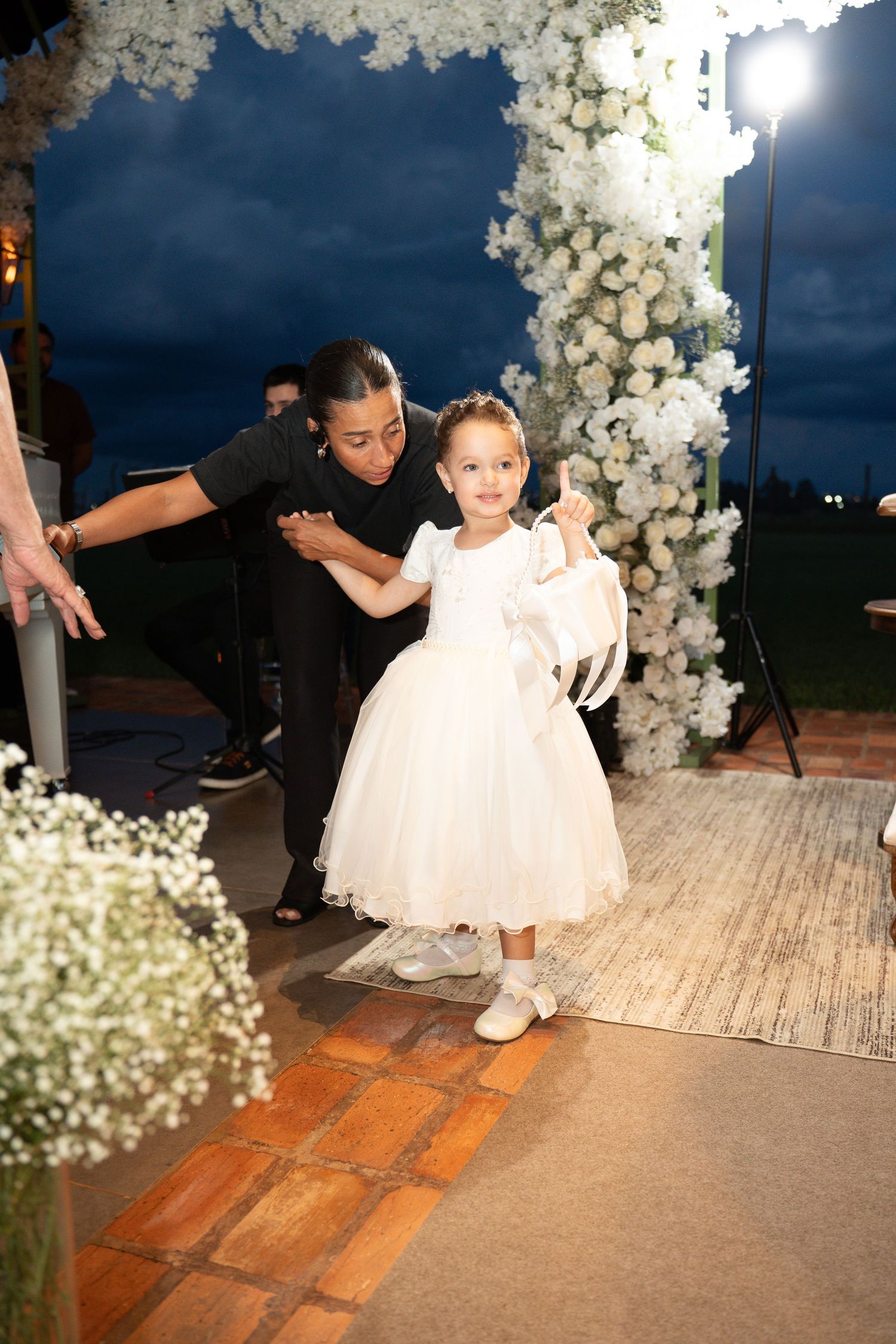 A young flower girl in a white dress walks down the aisle at an outdoor evening wedding ceremony.