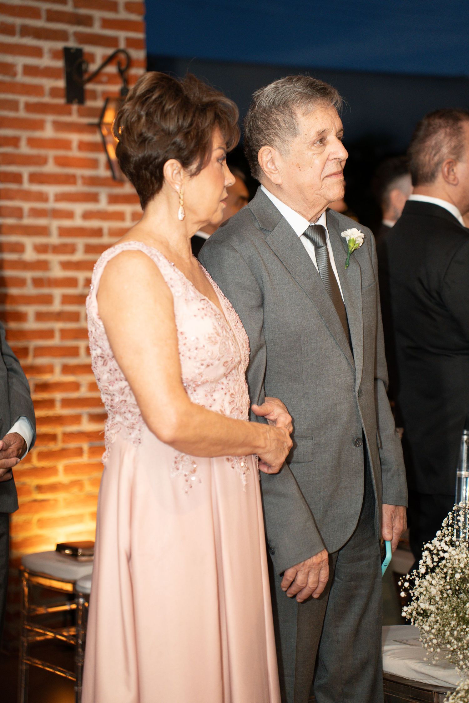Elderly couple dressed formally at a wedding, woman in pink gown and man in gray suit with boutonniere.
