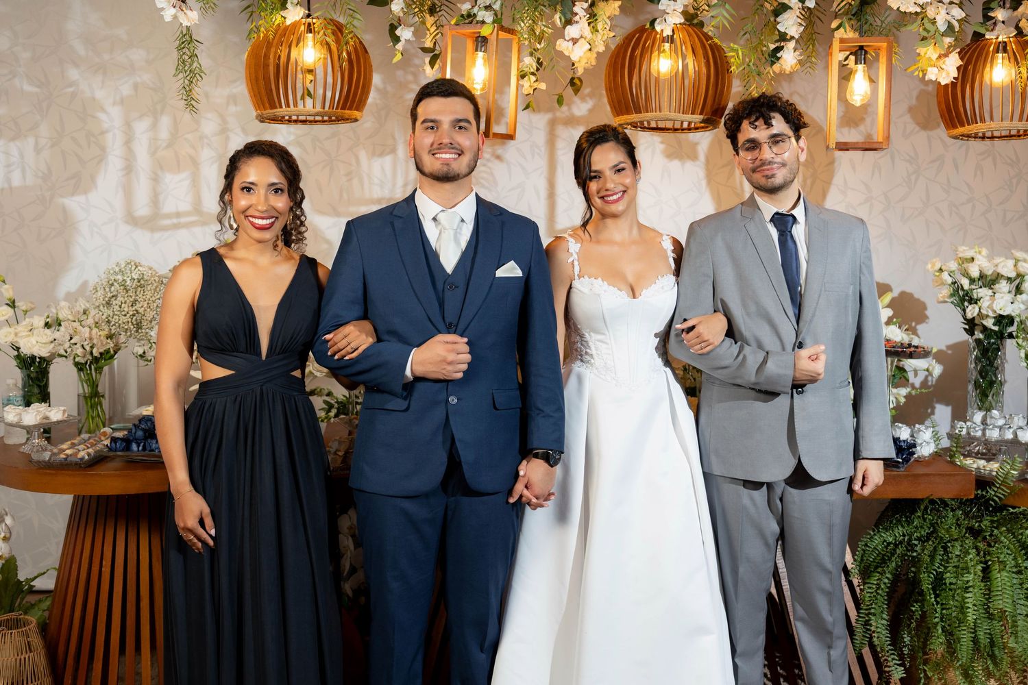 Wedding party of four posing elegantly, bride in white gown, groom in navy suit, guests in black dress and gray suit.