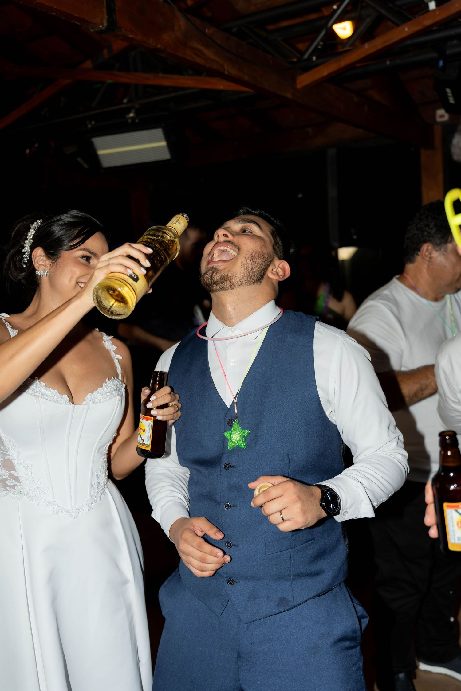 Bride pours drink into groom's mouth at wedding reception, both laughing and celebrating in festive venue.