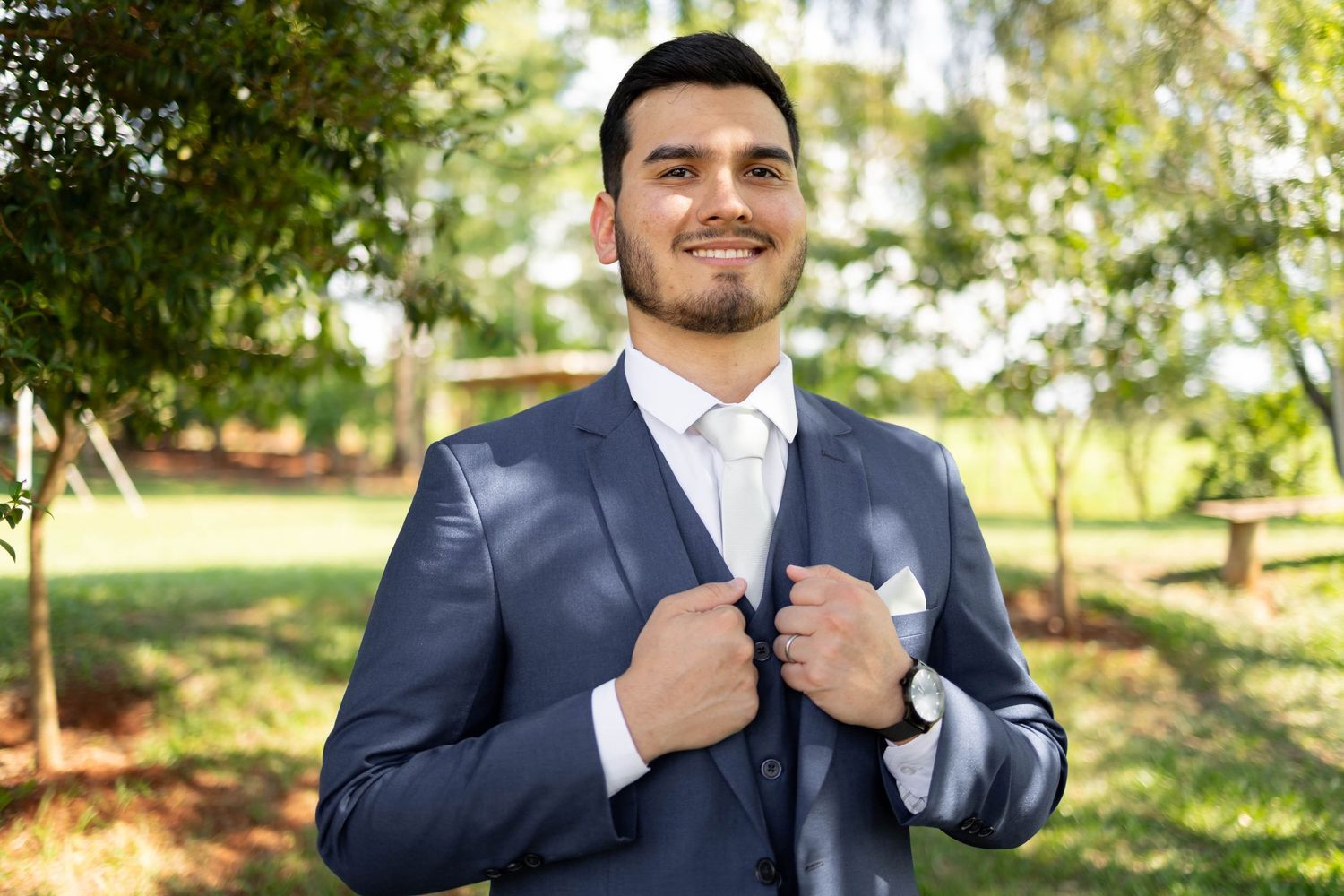 A well-dressed young man in a navy blue suit adjusts his jacket lapels while smiling outdoors among green trees.