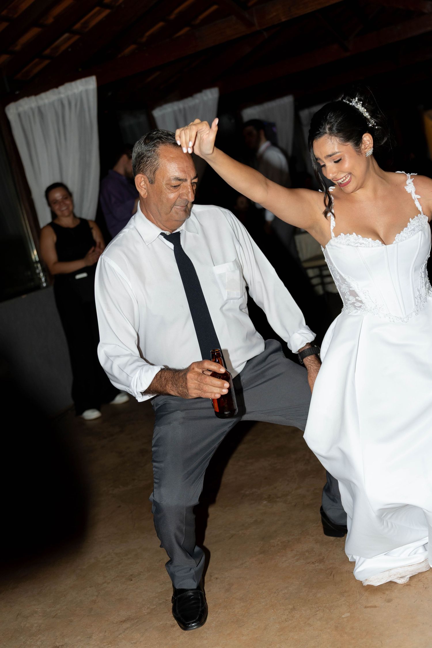 Bride in white gown dances joyfully with older man in white shirt and tie at wedding reception.