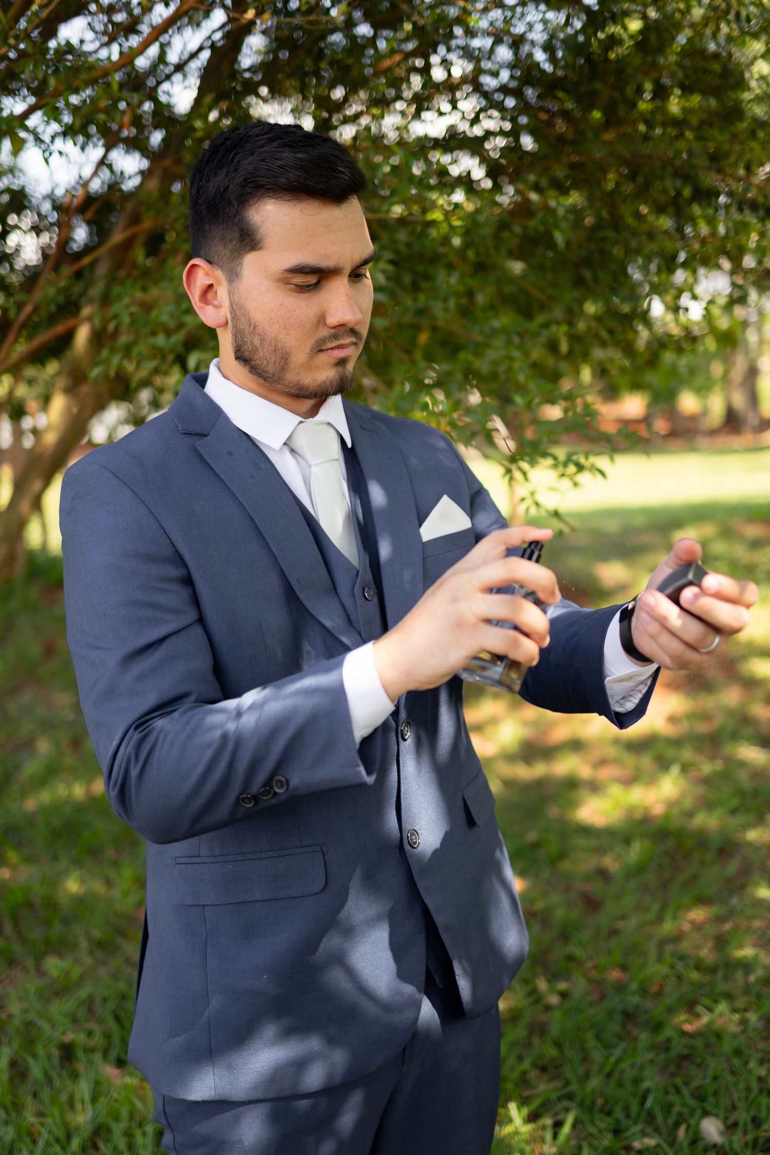 Groom in gray suit applying cologne outdoors before wedding ceremony.