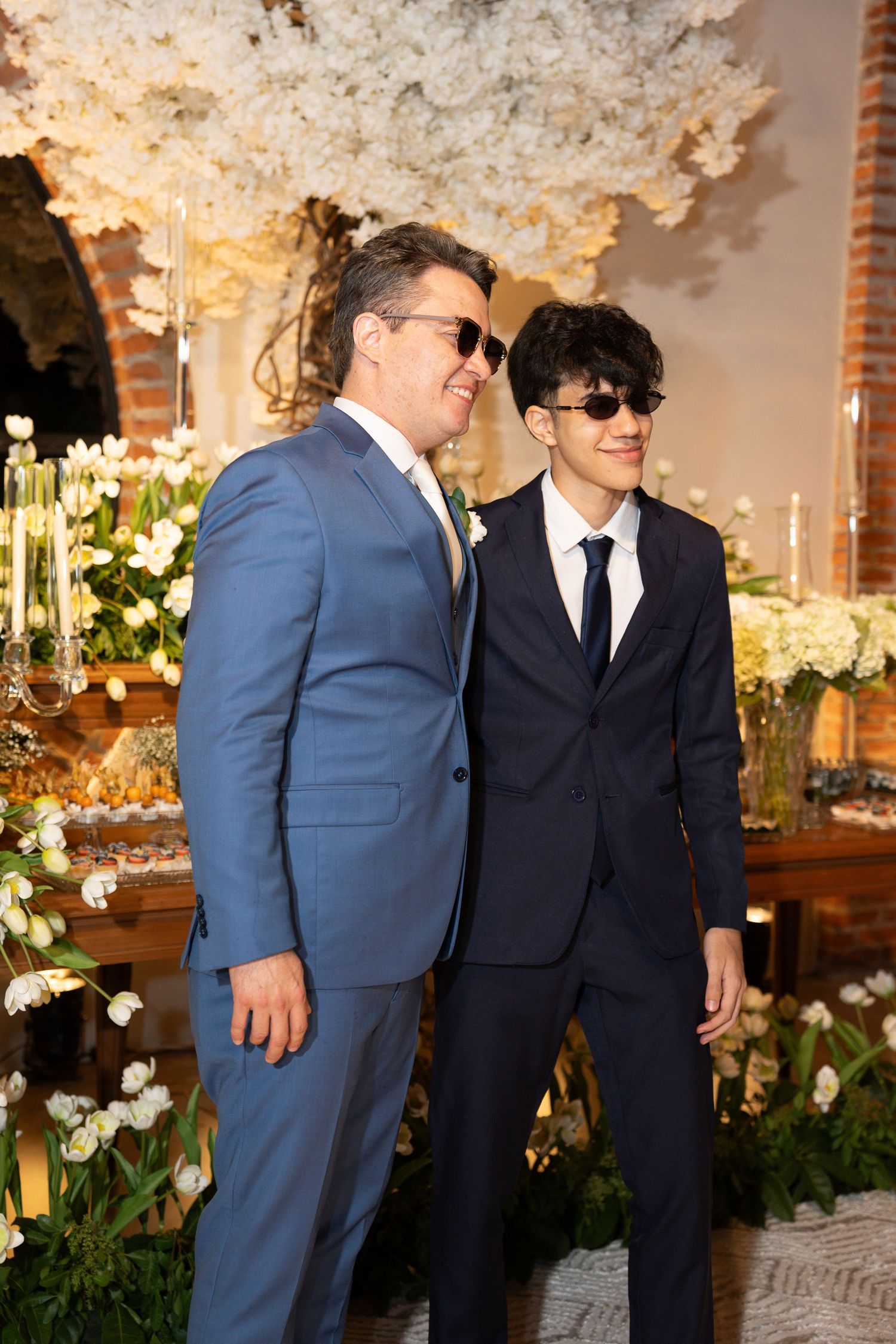 Two men in suits and sunglasses posing at an elegant wedding reception with white floral decorations.