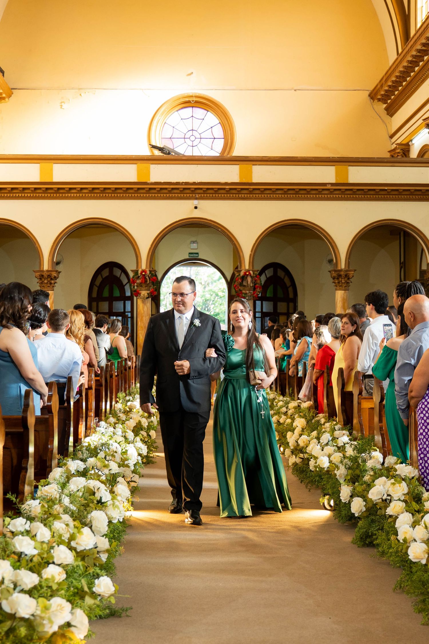A man in a suit escorts a woman in an emerald green gown down a flower-lined church aisle during a wedding ceremony.