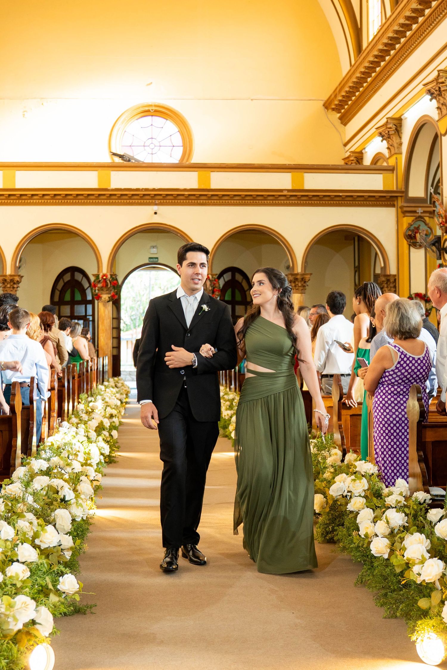 Couple walks down church aisle, man in black suit, woman in green gown, flanked by white floral arrangements.