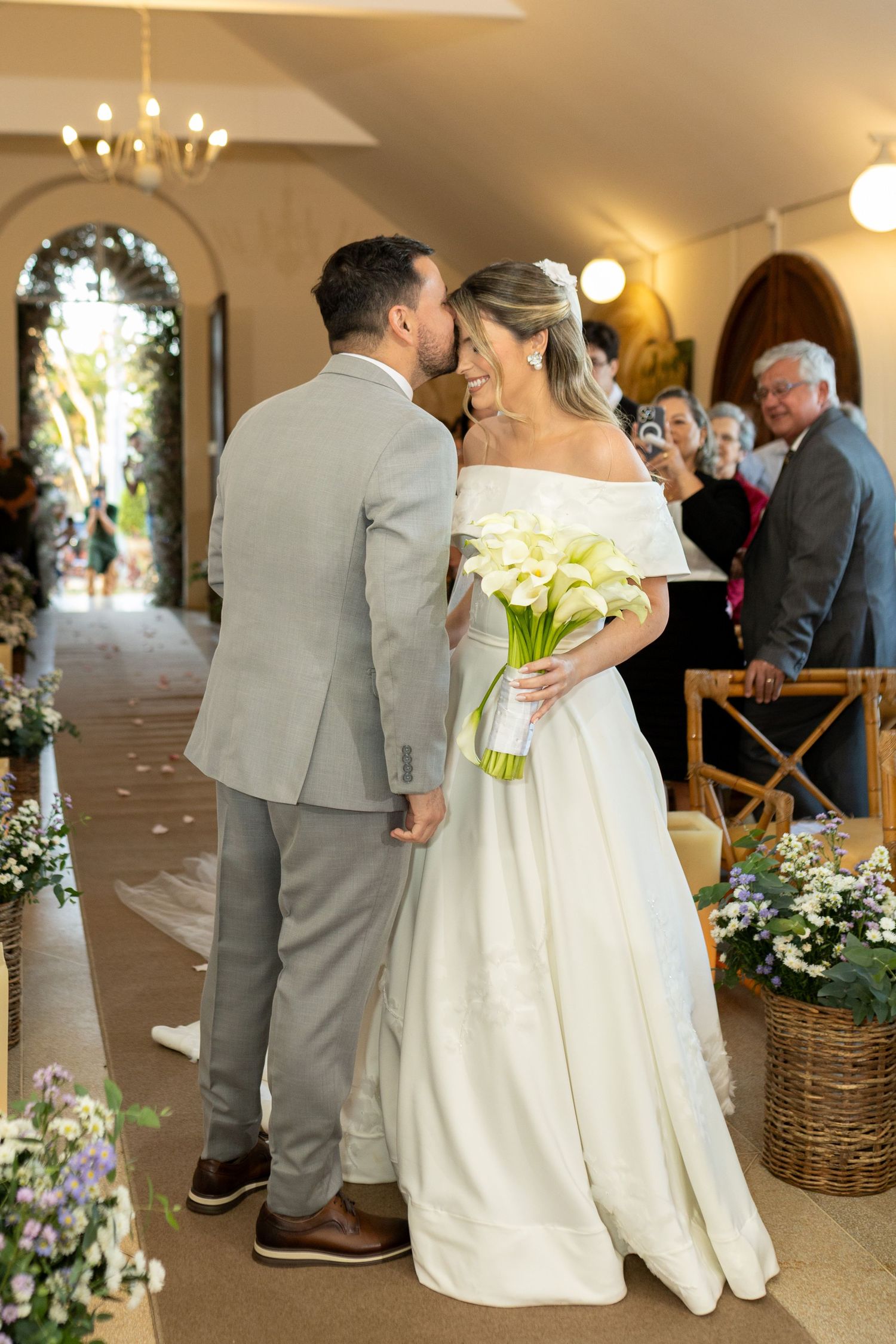 Bride and groom share a kiss at the altar, bride holding white calla lily bouquet in elegant off-shoulder gown.