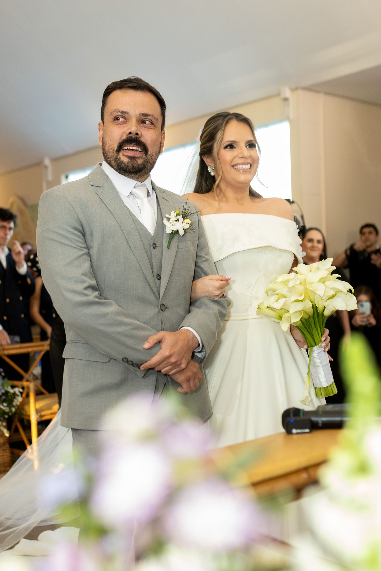 Smiling bride holding white calla lilies bouquet stands with groom in gray suit at church wedding ceremony altar.