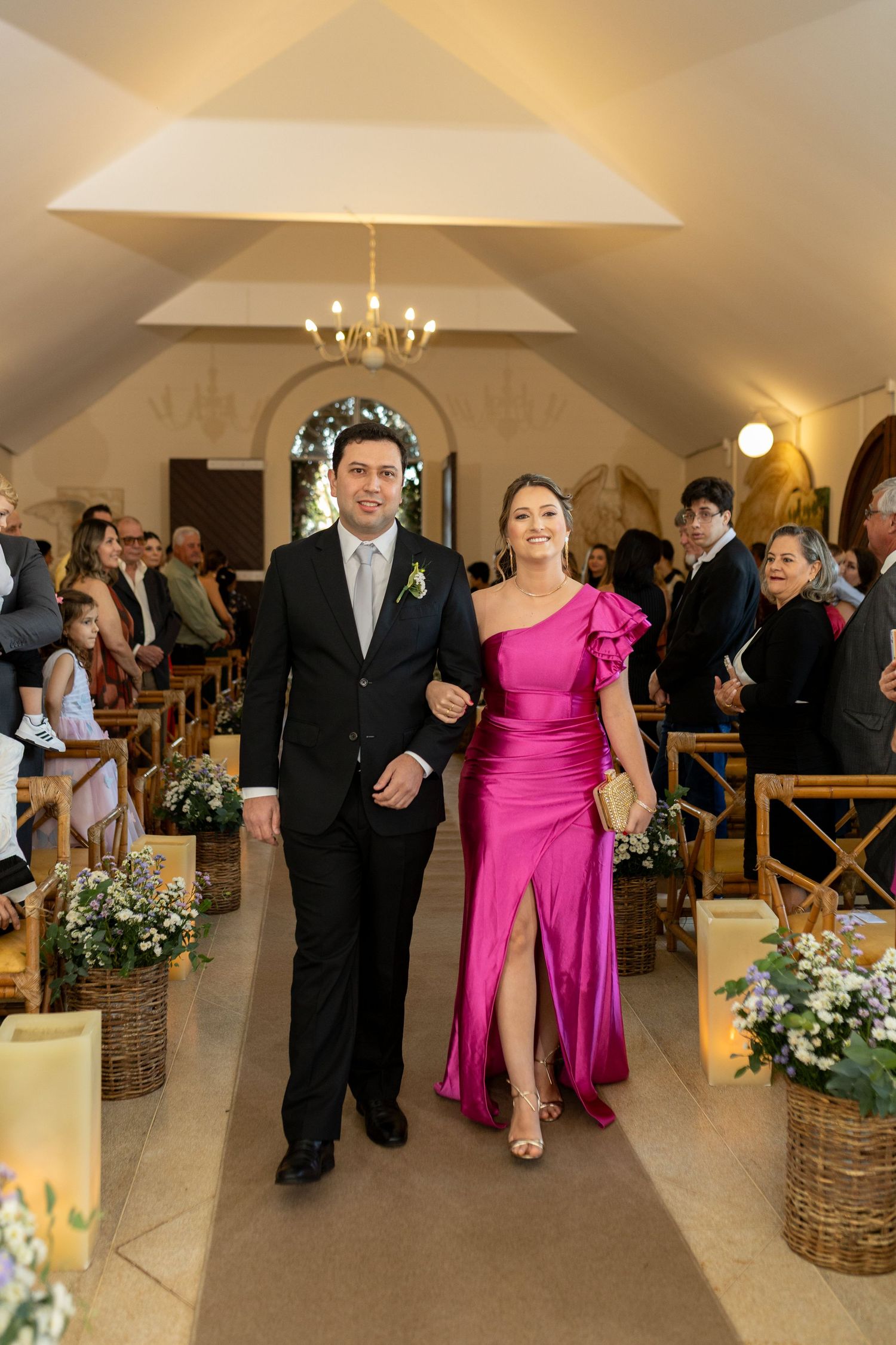 A couple walks down the aisle of an elegant chapel, man in black suit, woman in pink satin gown.