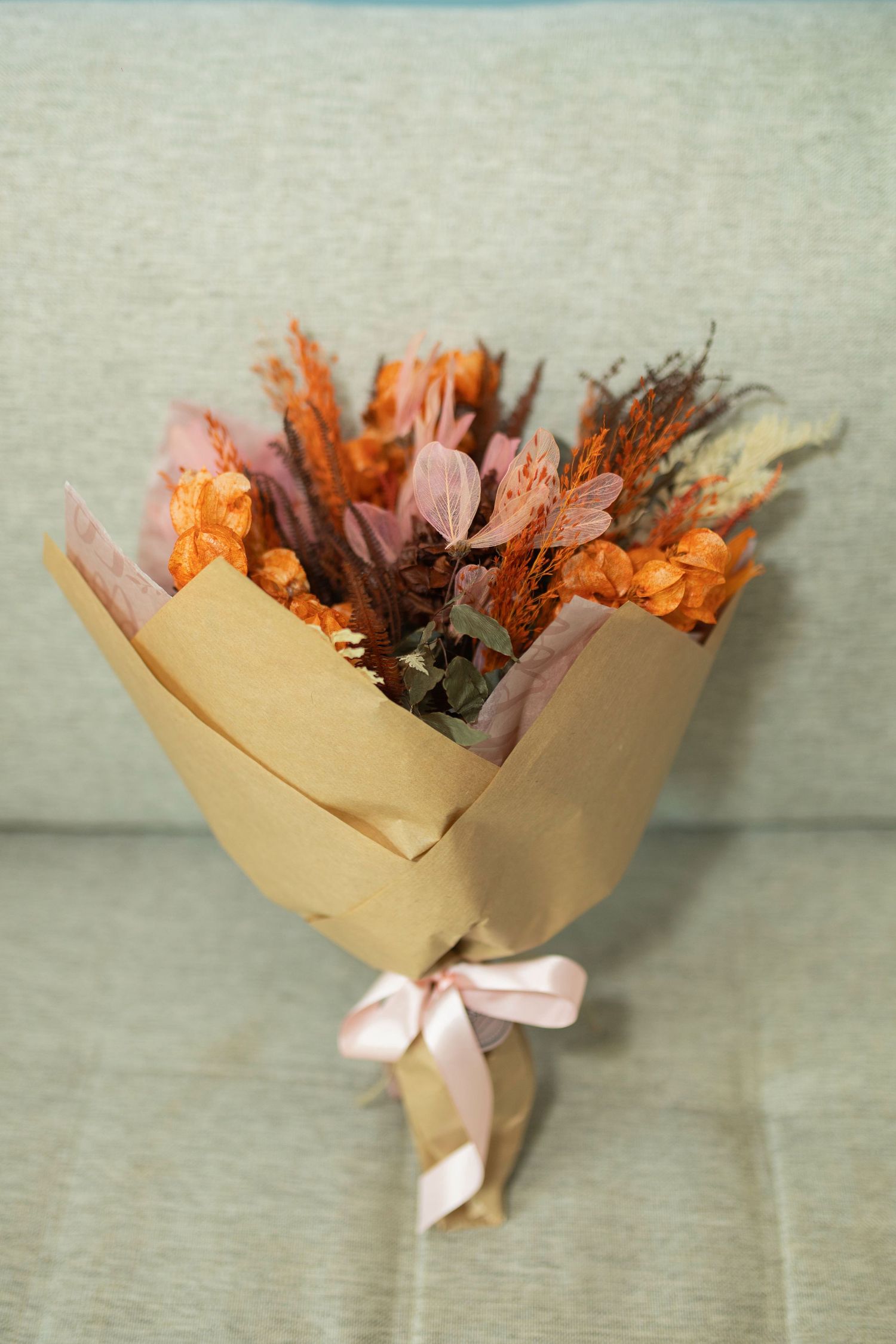Dried flower bouquet in kraft paper wrap with pink ribbon, featuring orange and pink blooms on a gray sofa.