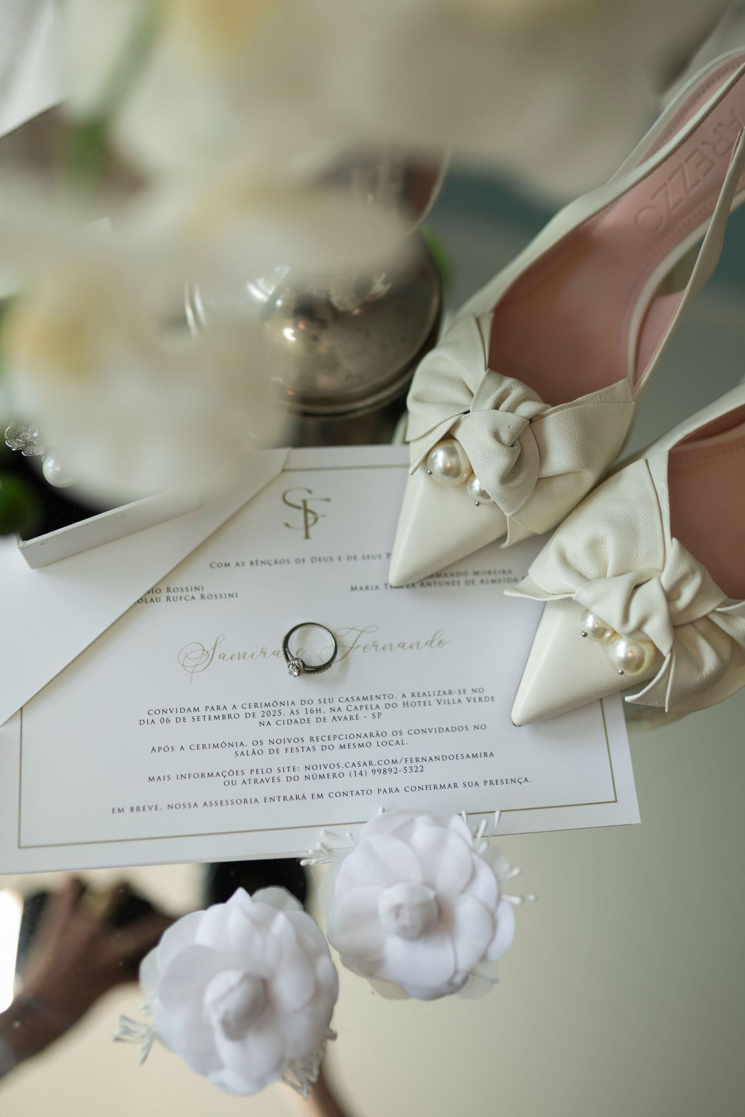 Elegant wedding flat lay with ivory bow heels, pearl accents, floral invitation, engagement ring, and white flowers on white background.
