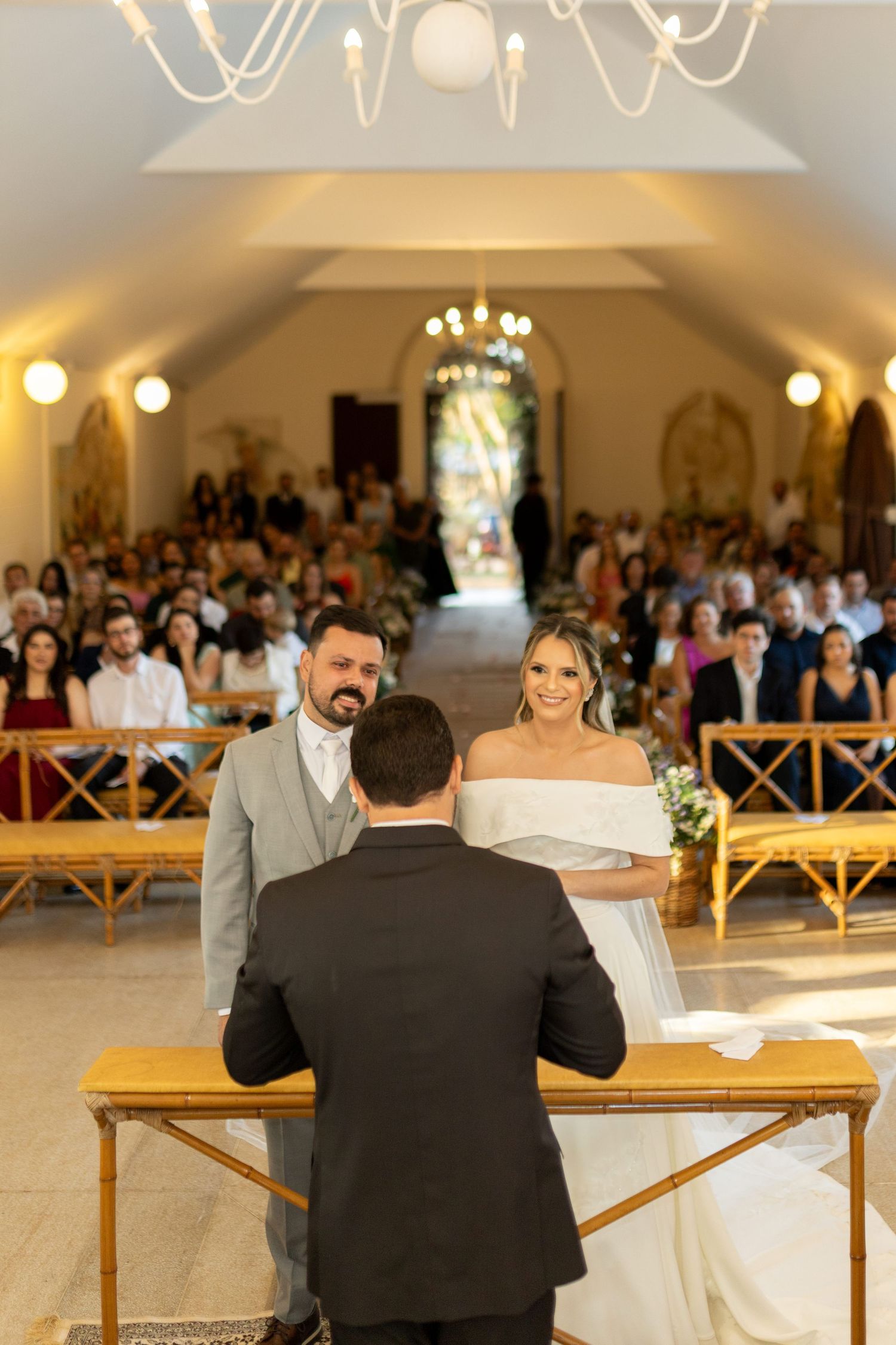 A smiling bride in a white off-shoulder gown and groom in gray suit stand at the altar during a wedding ceremony.