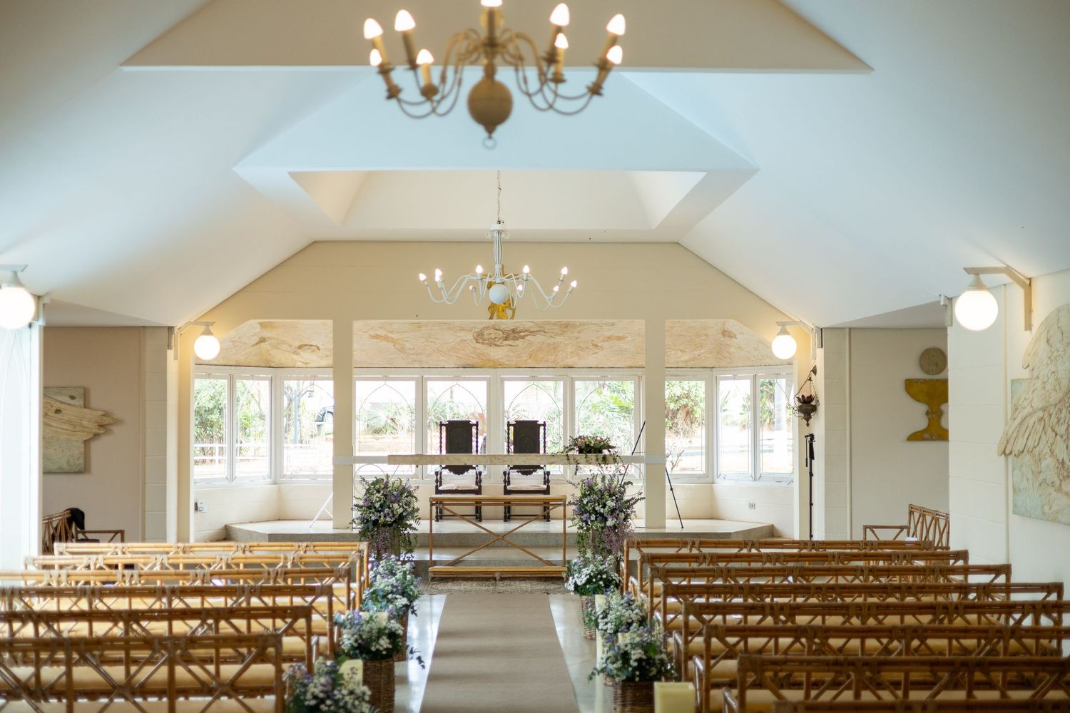 Elegant wedding ceremony venue with wooden pews, floral arrangements, chandelier lighting, and altar framed by large windows.
