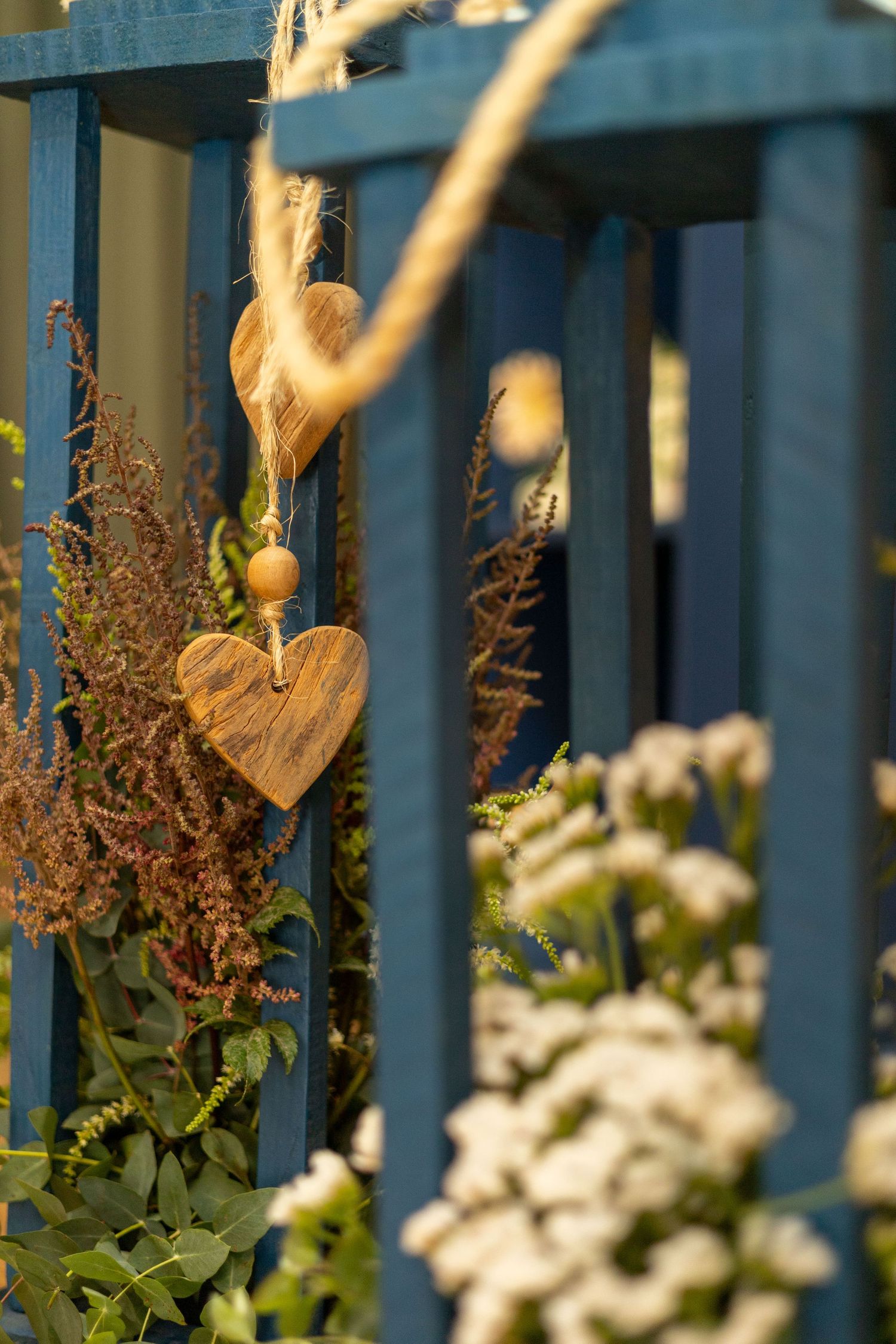 Wooden heart ornament hanging on a blue fence surrounded by lush green plants and white flowers in a garden.