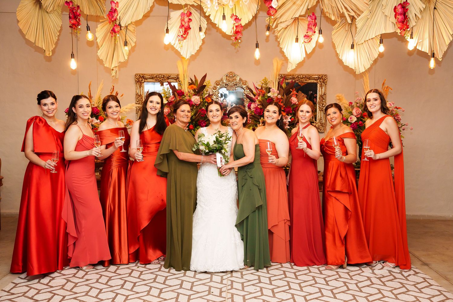 Bride in white gown poses with bridesmaids in red and olive green dresses at elegant floral wedding reception.