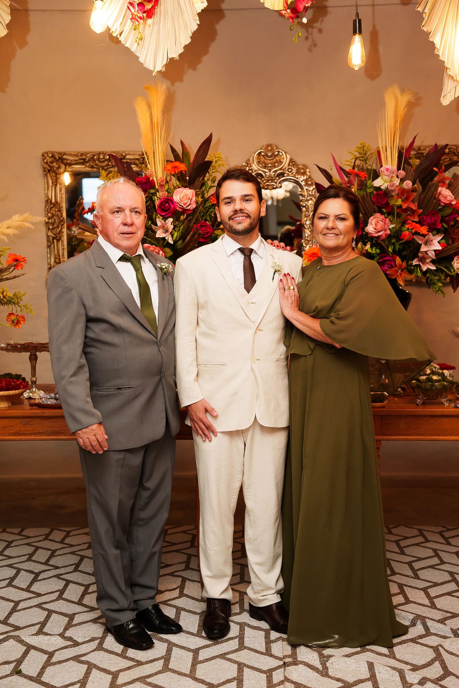 Groom in white suit poses with parents at floral wedding venue.